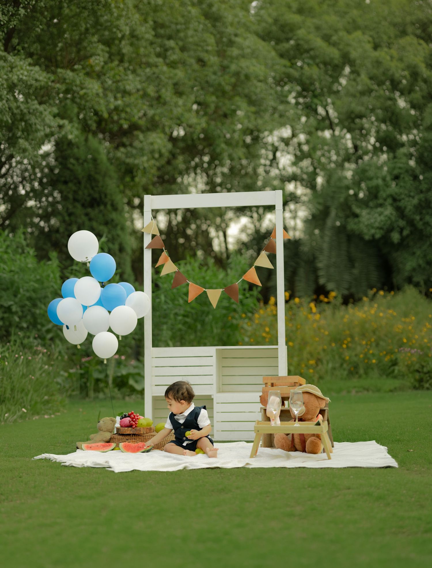 A toddler sits on a white blanket outdoors at a birthday setup with blue balloons, bunting, and a small wooden chair.