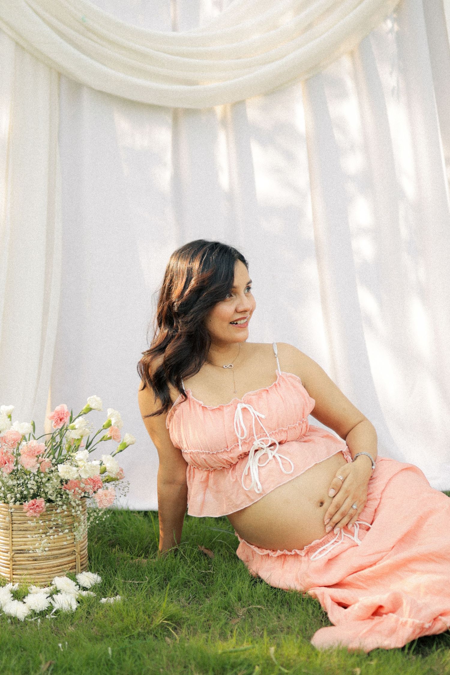 Smiling pregnant woman in pink crop top and skirt sitting on grass near flowers with white curtain backdrop.