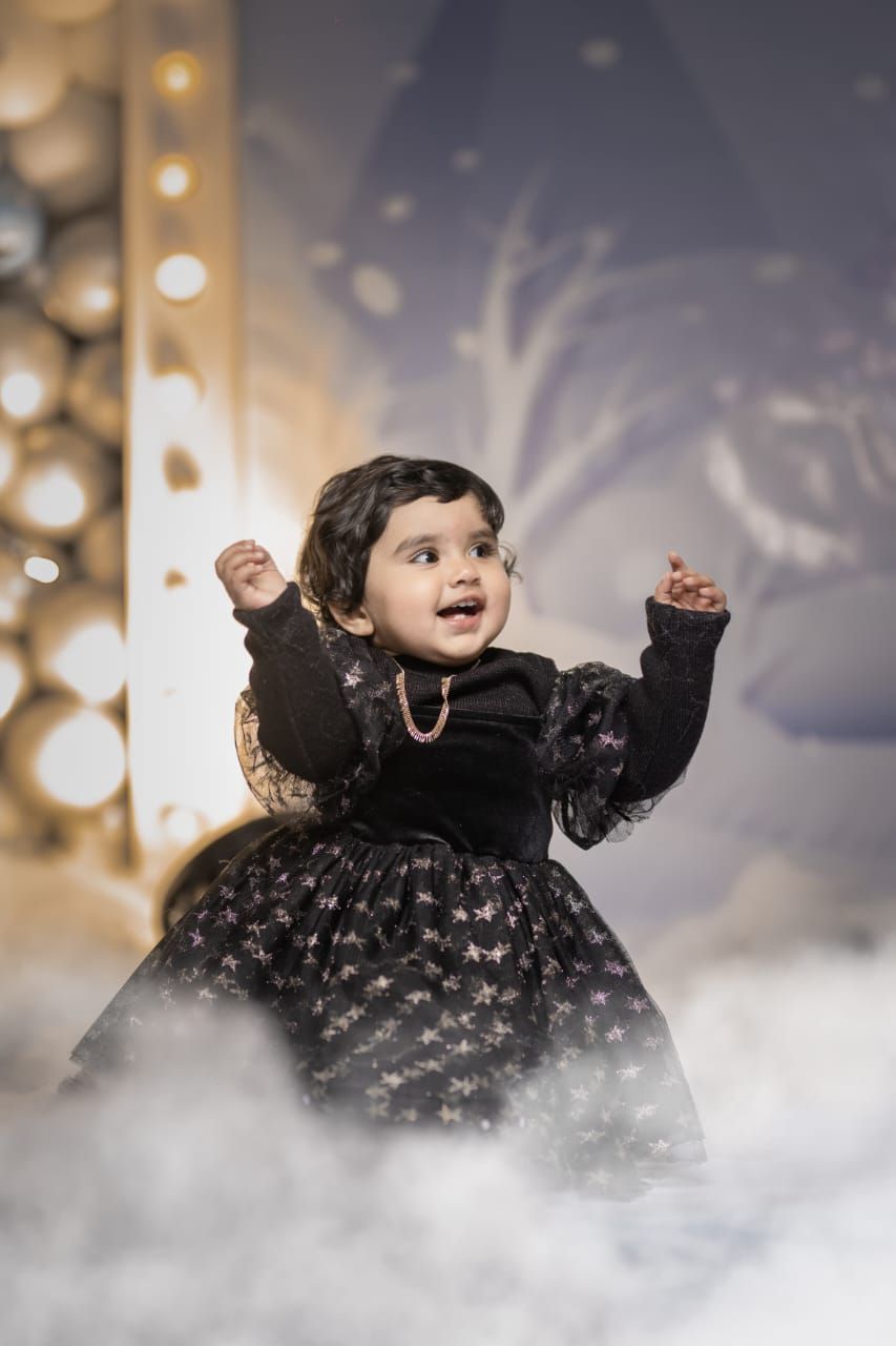 A joyful toddler in a black star-print dress with raised arms, posing in a dreamy bokeh-lit studio setting.