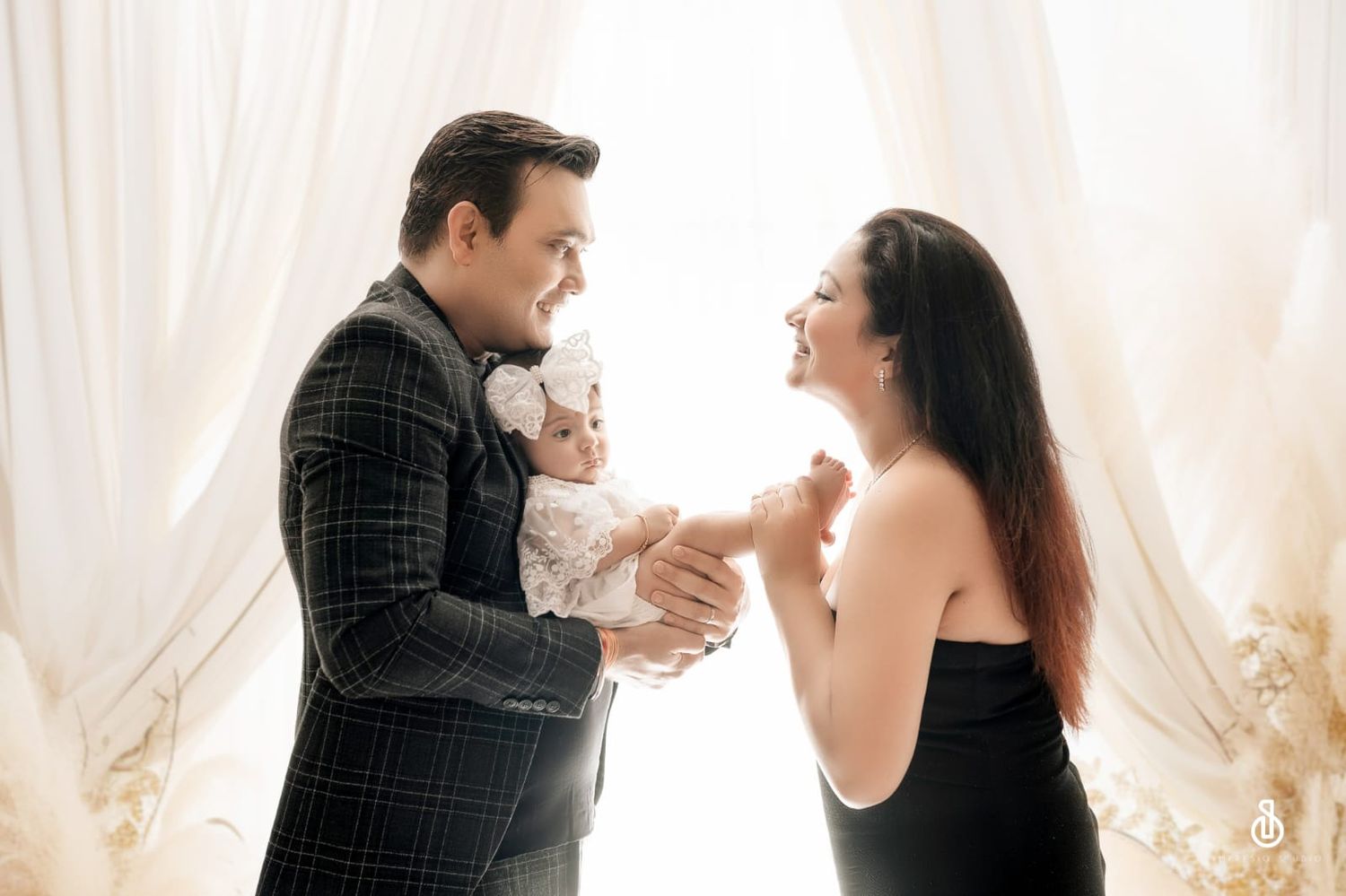 A couple in elegant attire shares a joyful moment with their baby in a softly lit, curtained studio setting.