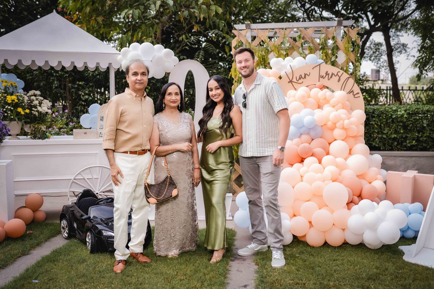 Four elegantly dressed guests pose together at a pastel balloon-decorated outdoor celebration party.