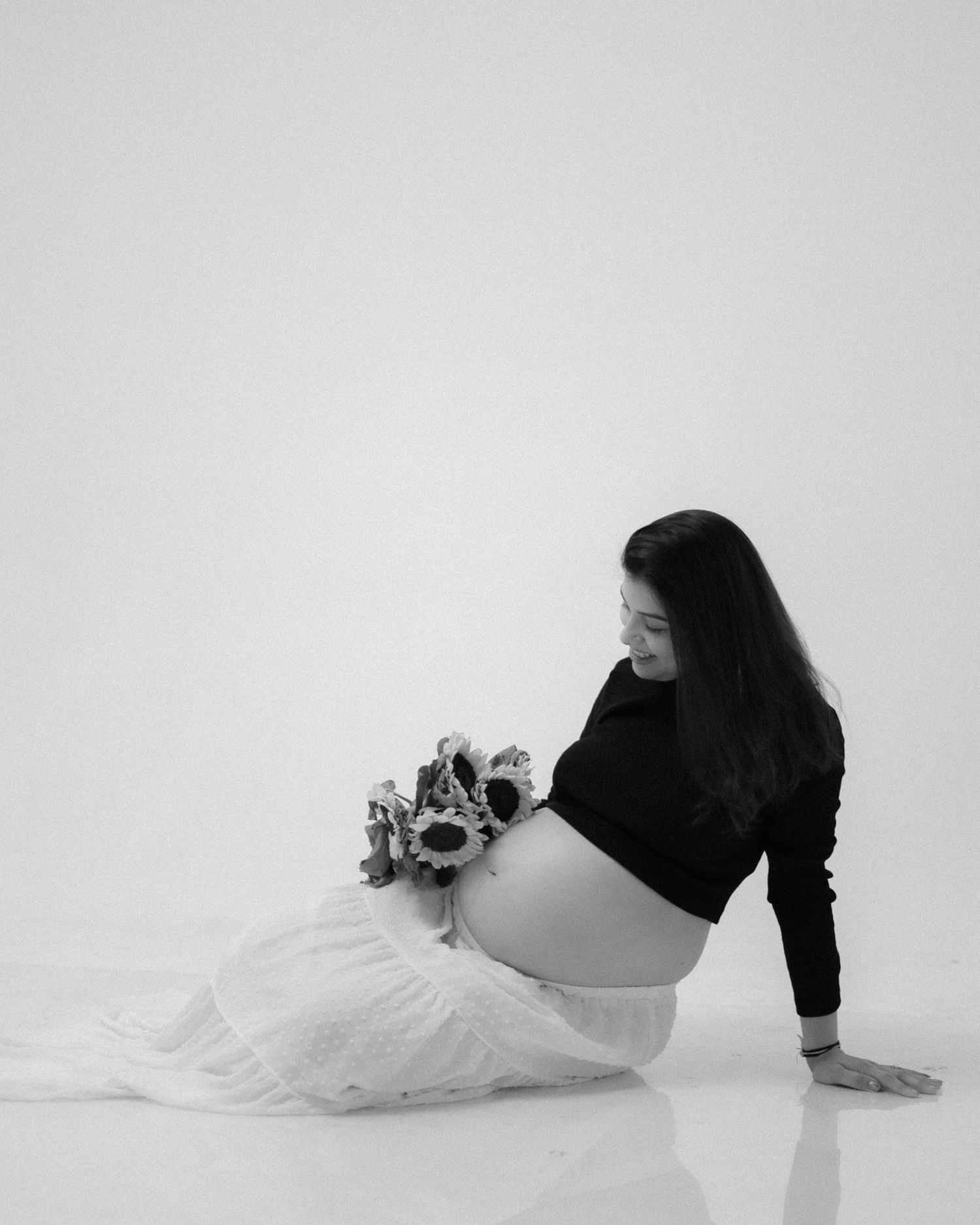 Black and white maternity photo of a woman sitting on the floor holding flowers against her baby bump.