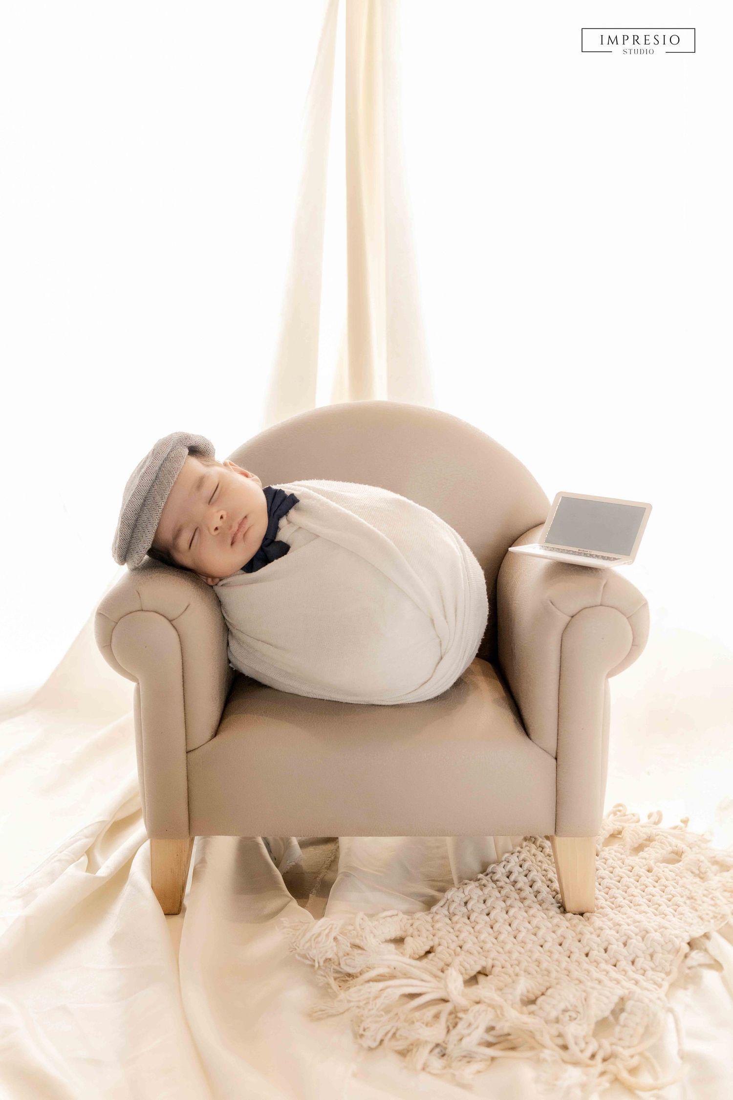 Sleeping newborn baby swaddled in white wrap resting on beige armchair with flat cap and small frame prop.