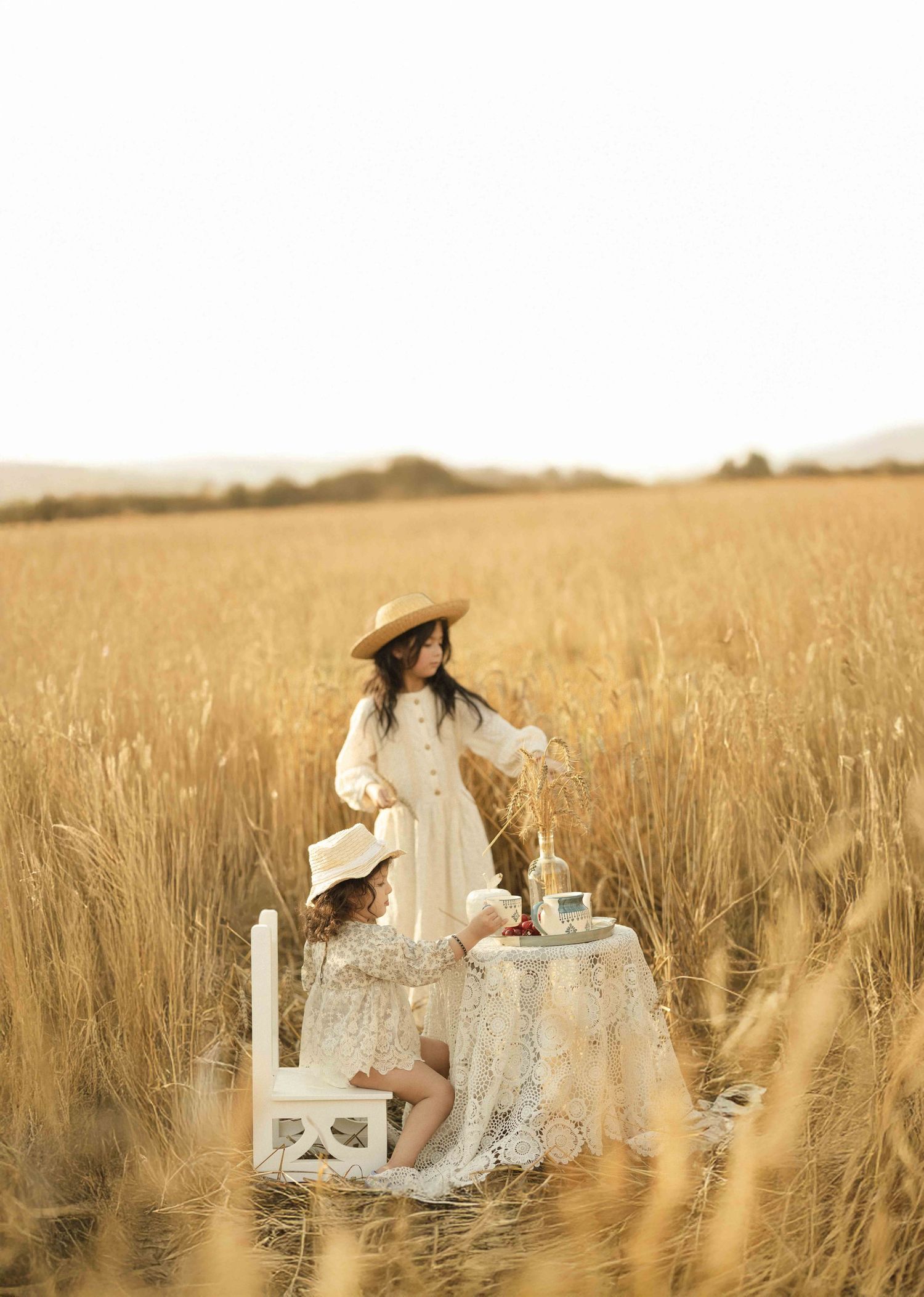 Two girls in white dresses enjoy a tea party in a golden wheat field, one wearing a straw hat.