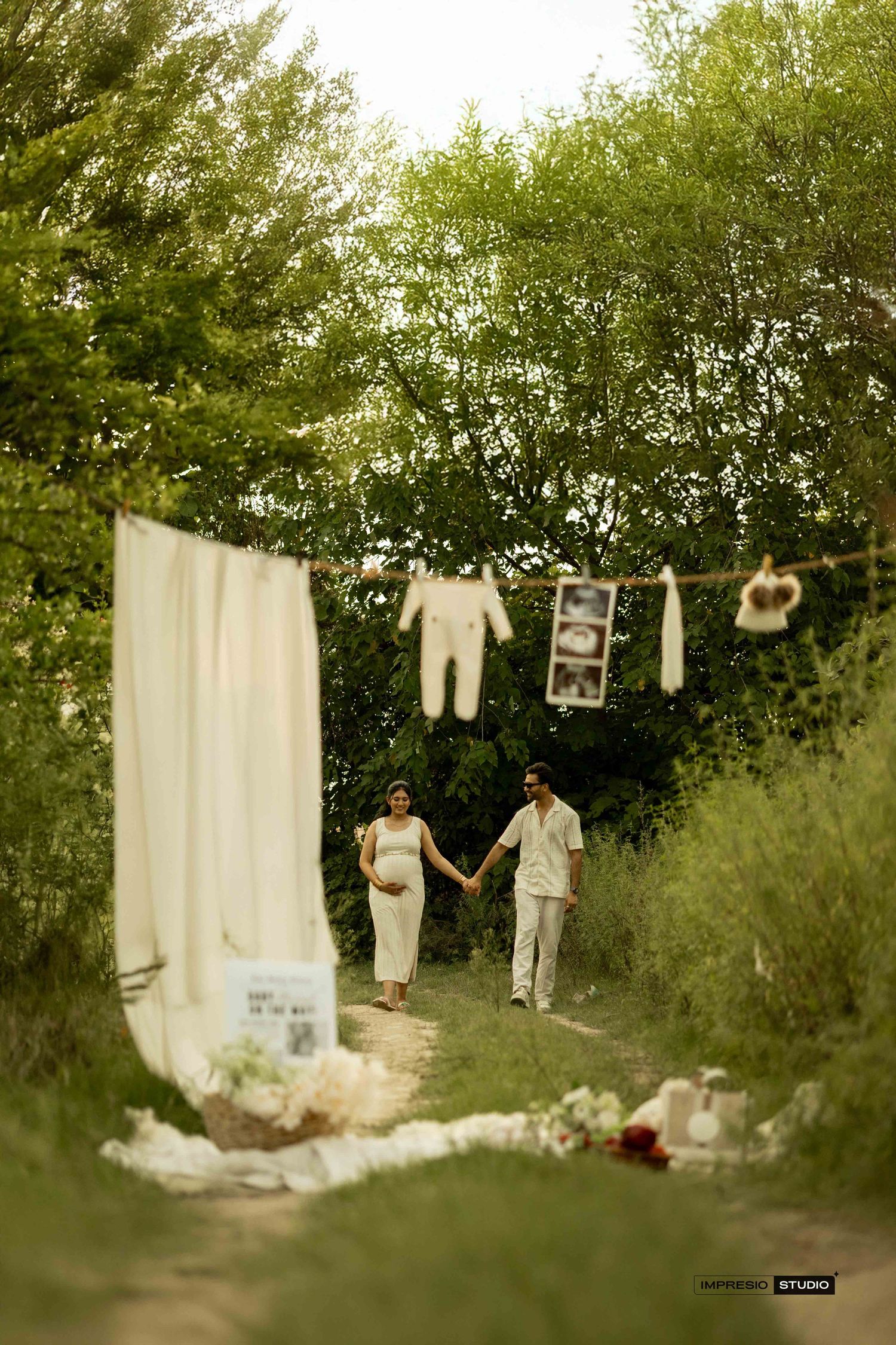 Couple holding hands during outdoor maternity photoshoot with baby clothes hanging on a line surrounded by lush greenery.