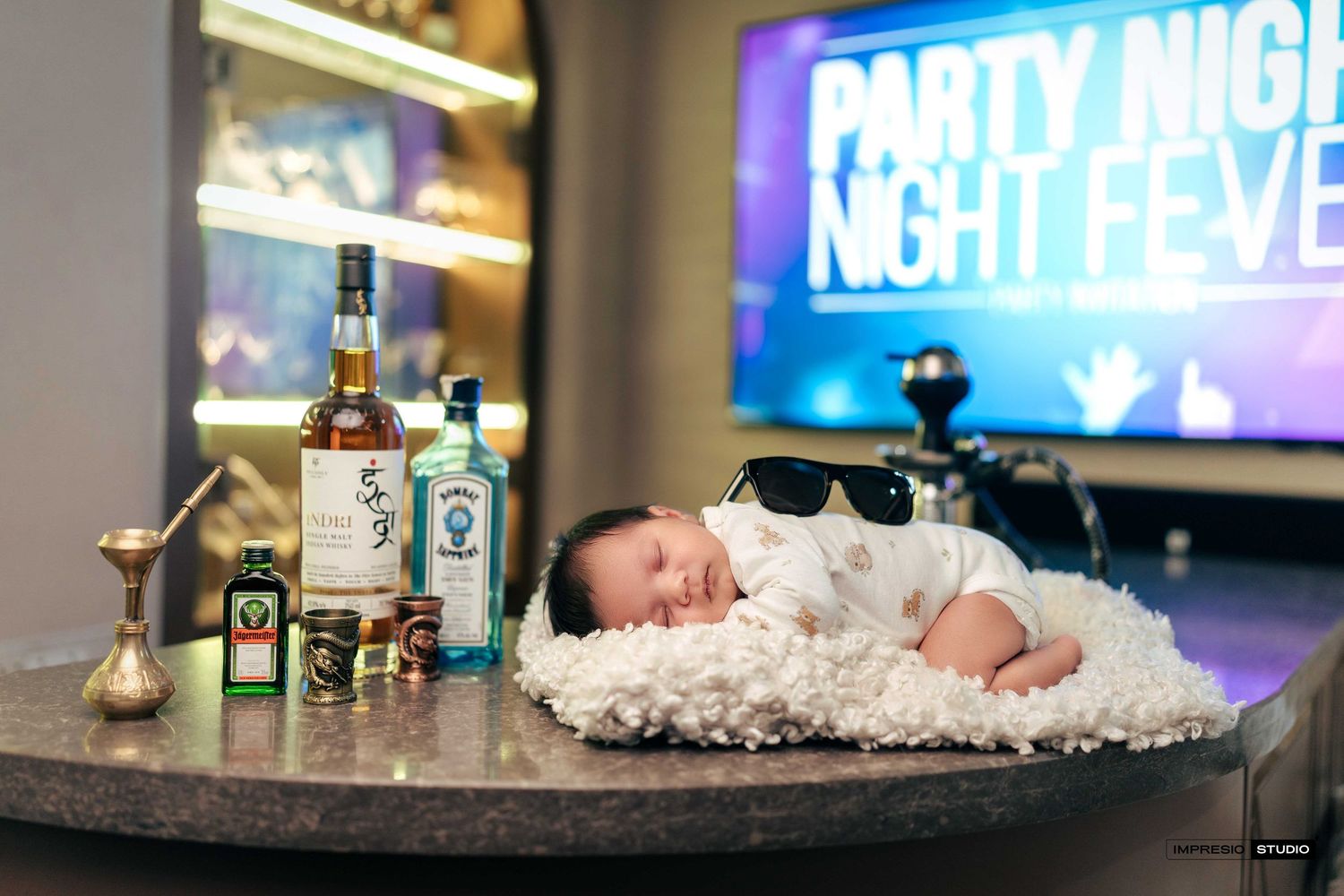 Newborn baby sleeping on fluffy white blanket surrounded by liquor bottles wearing sunglasses at a party-themed photo shoot.