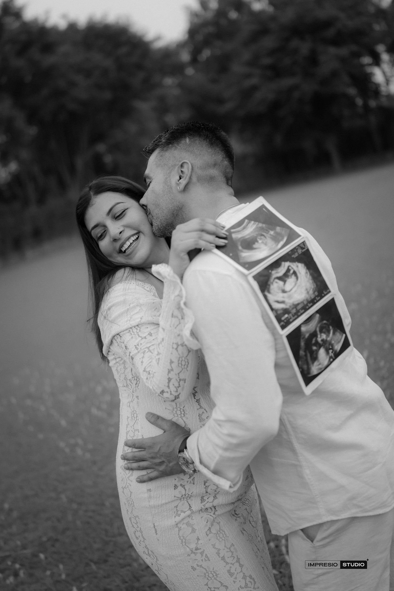 Black and white photo of a couple embracing outdoors, man kissing woman's cheek while holding ultrasound photos.