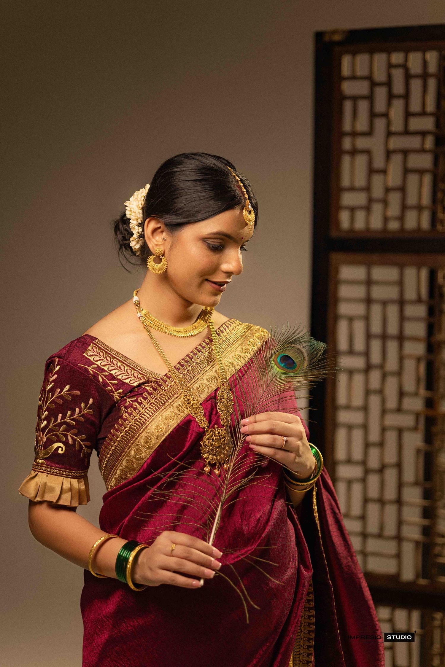A woman in a red silk saree with gold jewelry holds a peacock feather, dressed in traditional South Indian bridal attire.