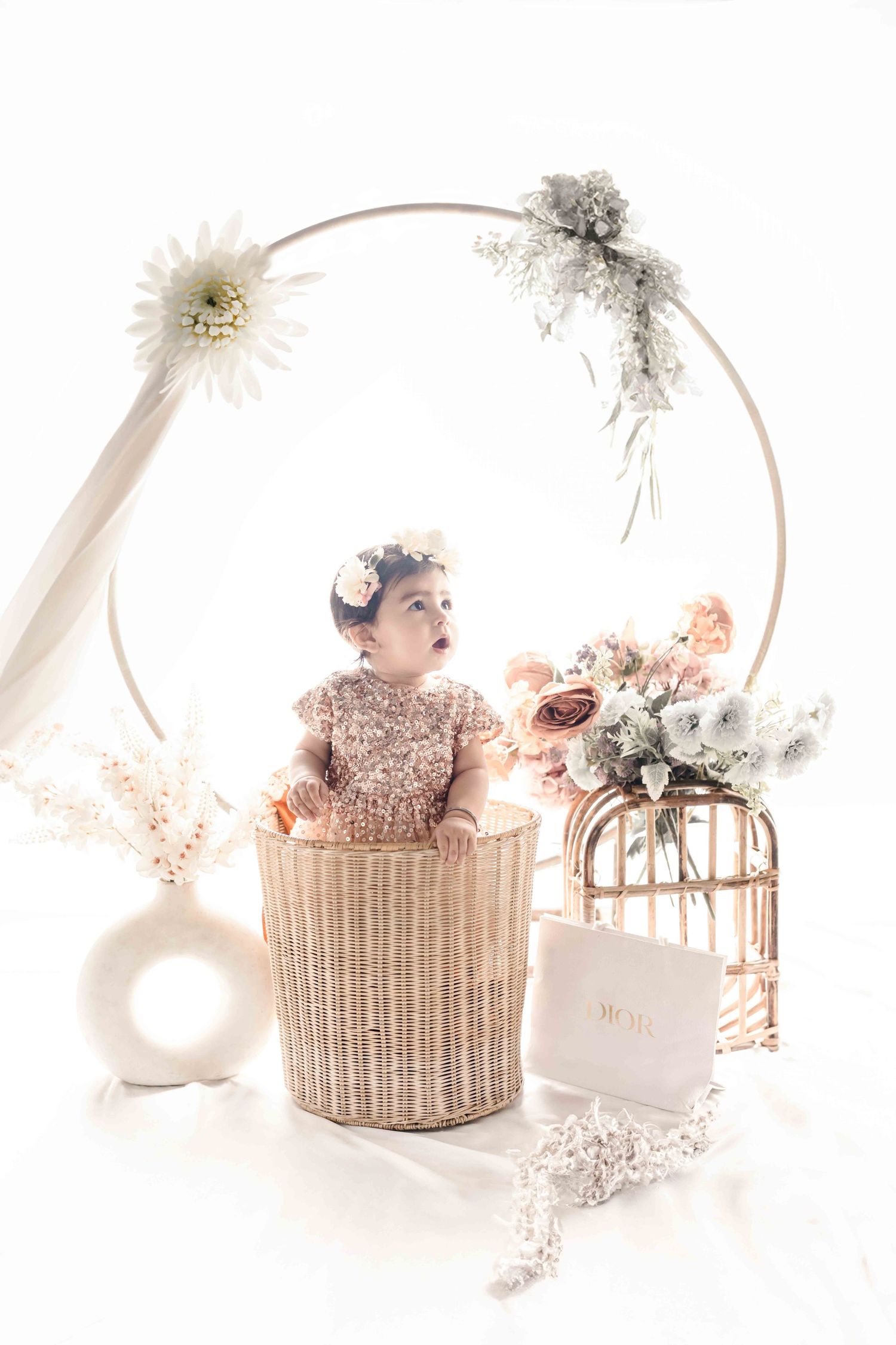 Baby girl in sparkly dress sits in wicker basket surrounded by flowers and circular floral hoop backdrop.