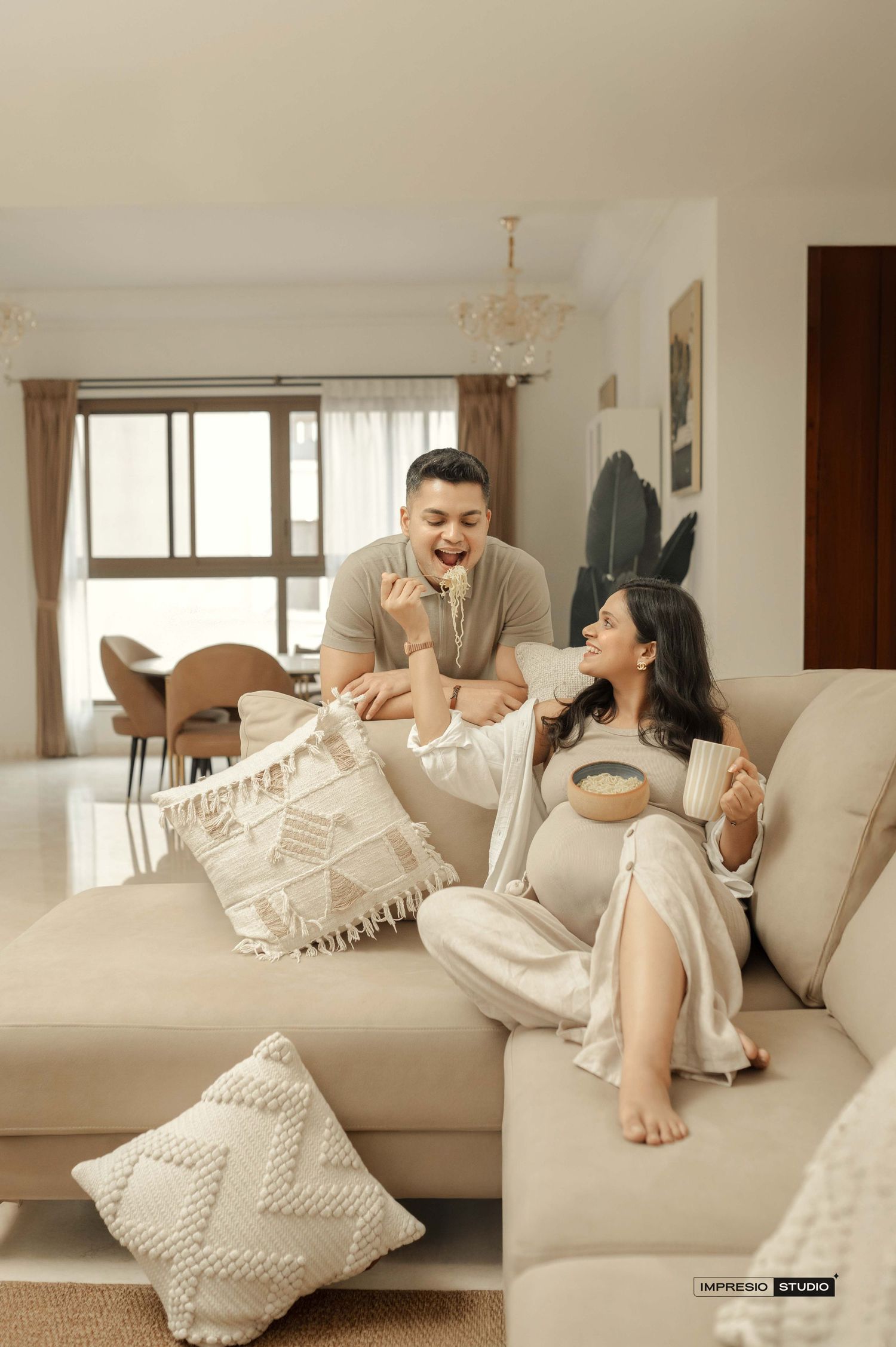 A couple laughs together on a beige sofa, woman visibly pregnant, sharing a joyful moment in a modern living room.