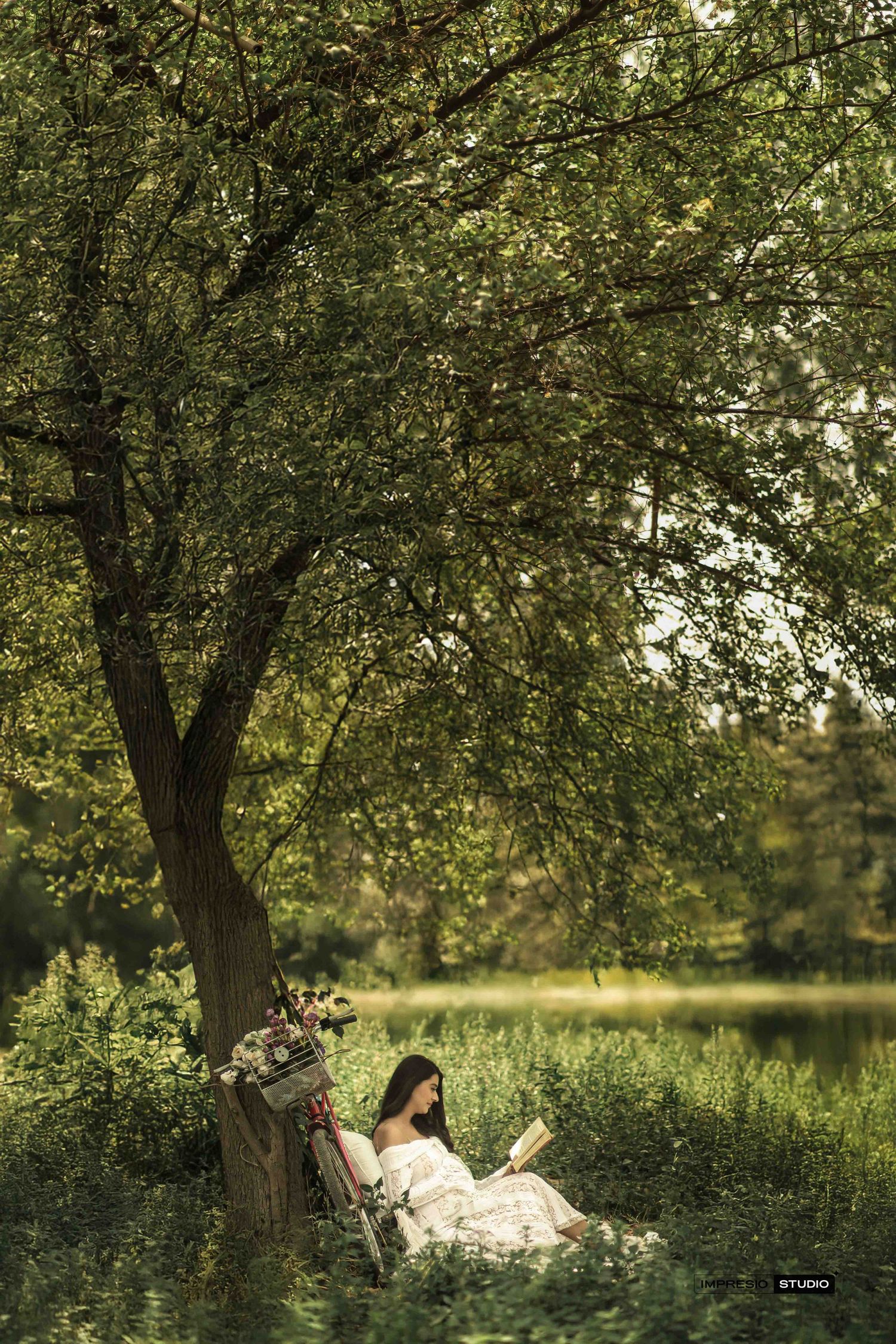 Woman in white dress reading a book under a large tree by a serene lake surrounded by lush green foliage.