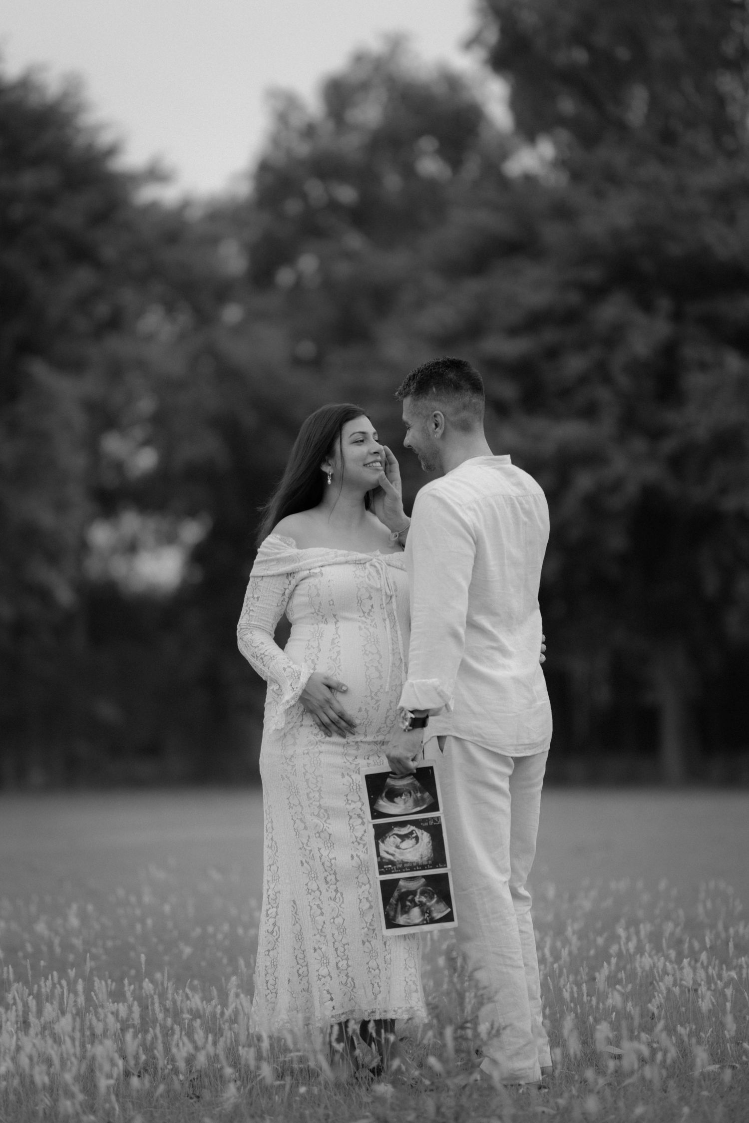 Black and white photo of a pregnant couple holding ultrasound images in a field, sharing a tender moment.