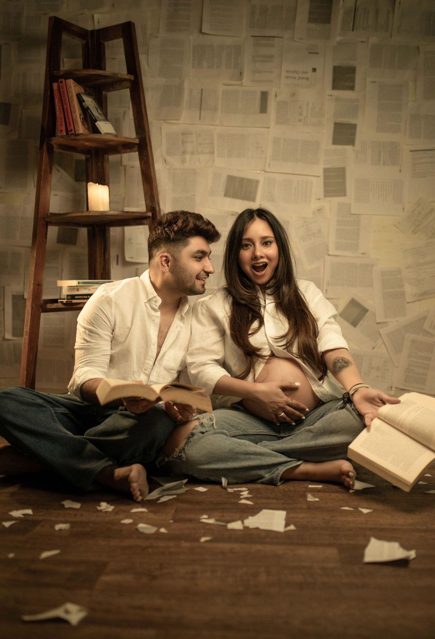 Pregnant couple sitting on floor reading books, surrounded by scattered pages and a wooden ladder shelf.