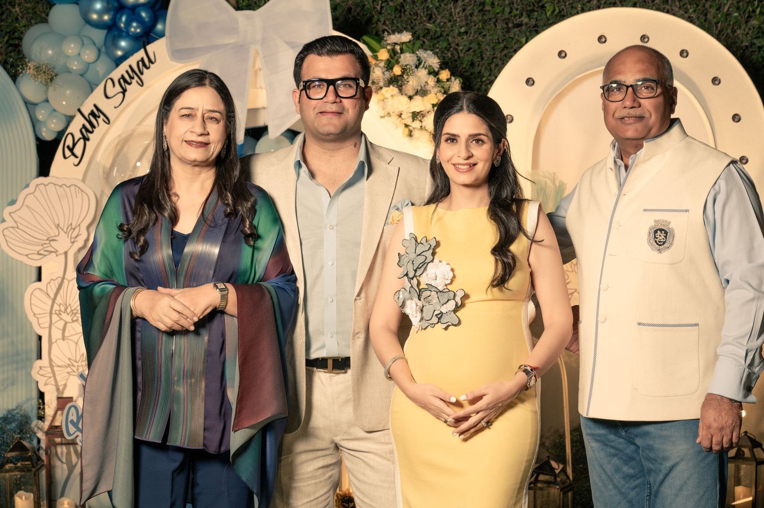 Four people posing at a baby shower event with blue and white decorations and a Baby Sanjay banner.