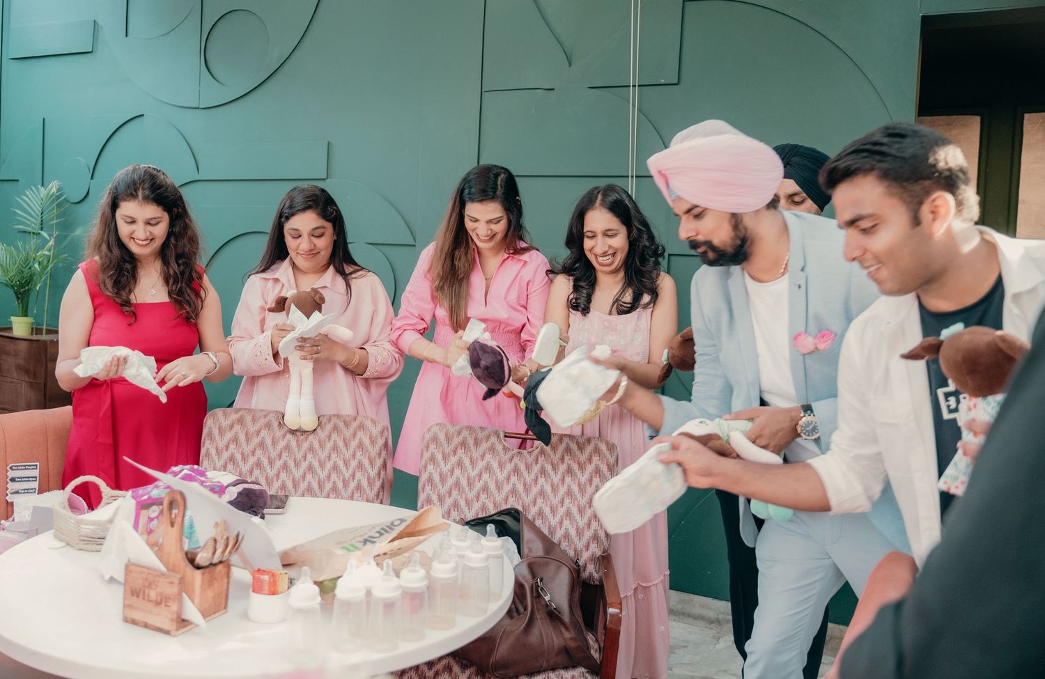 Group of people at a baby shower examining small baby items and gifts at a decorated table indoors.