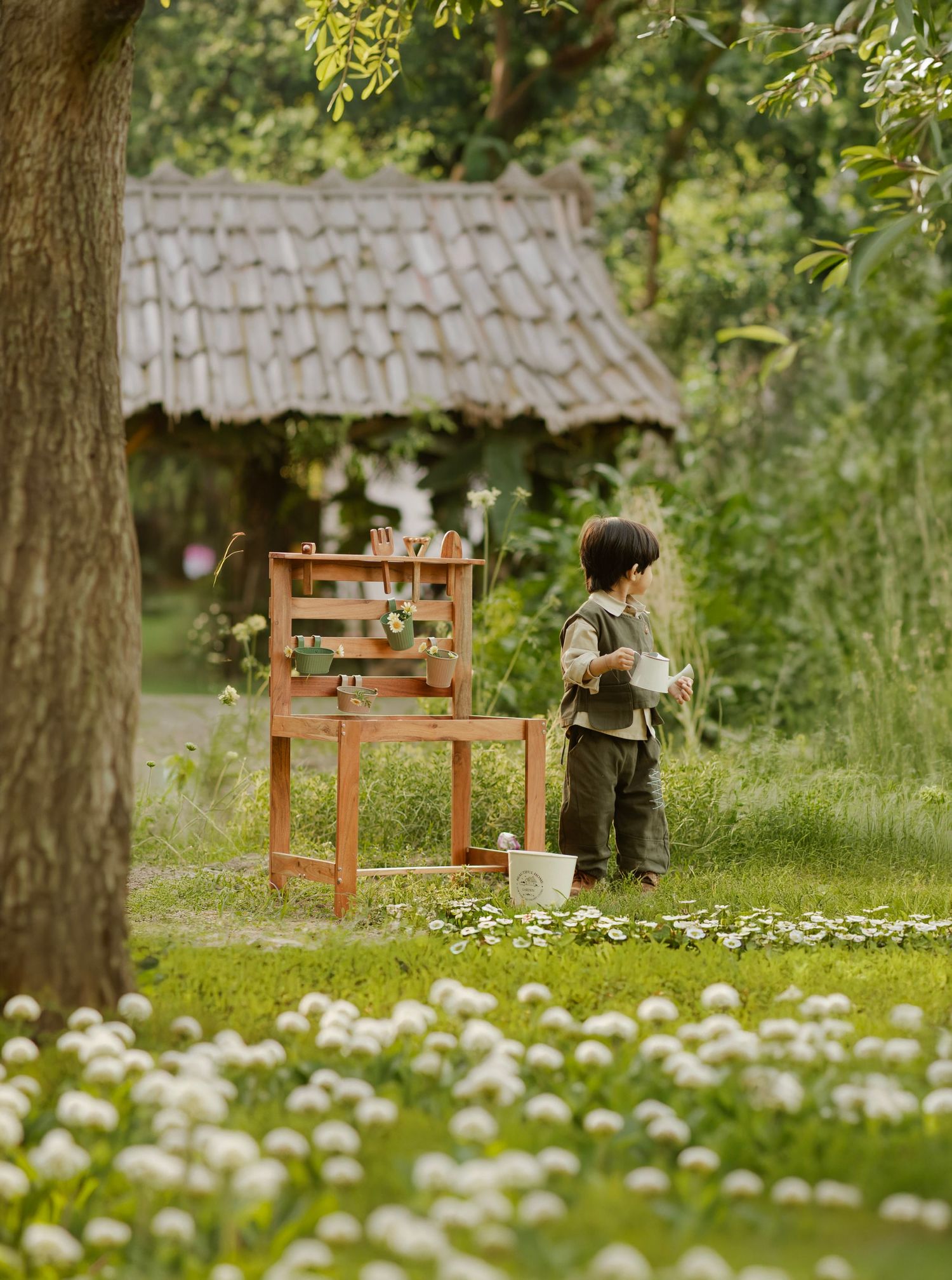 A young child waters plants in a lush garden beside a wooden chair and rustic thatched cottage.