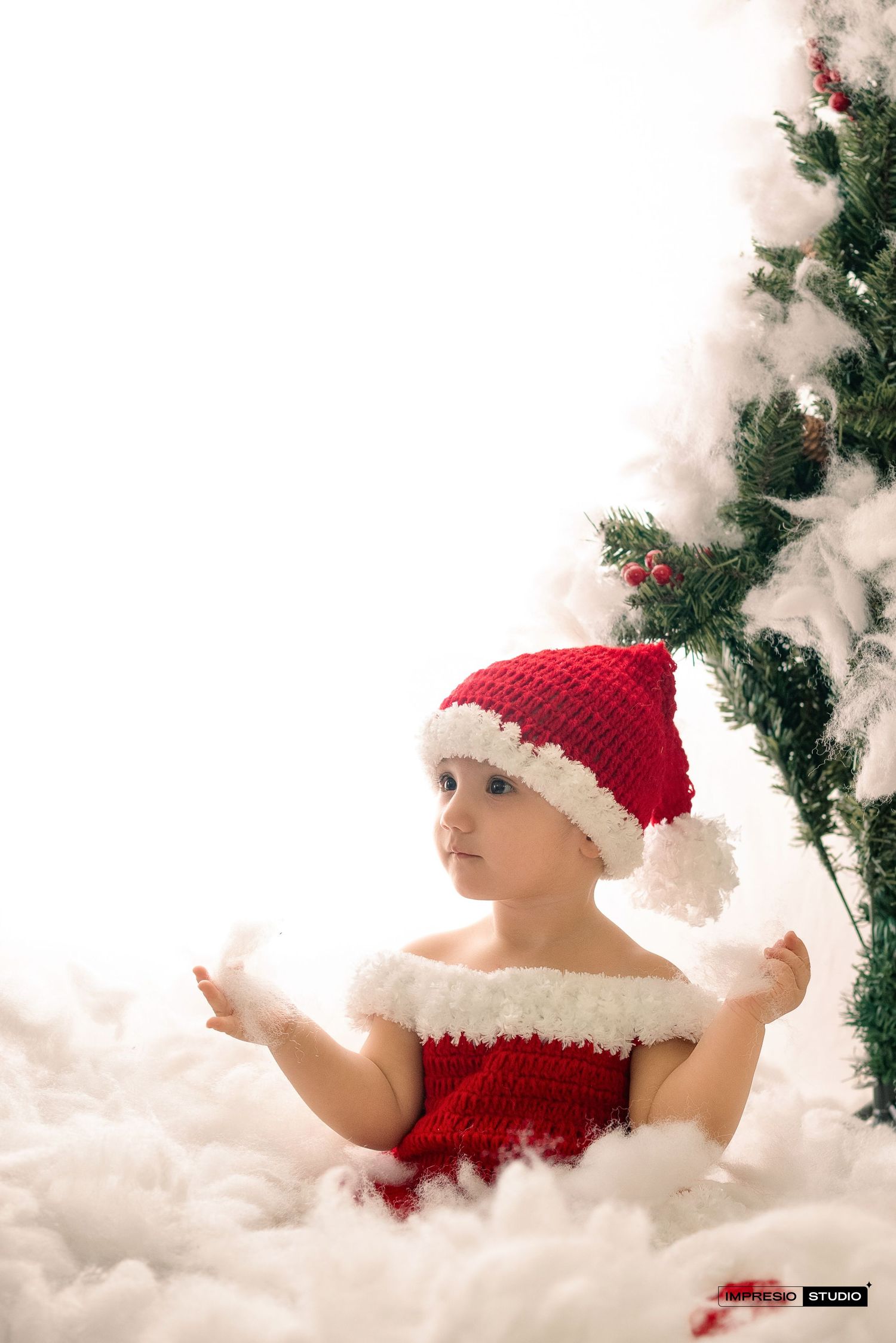 A small child wearing a red and white Santa outfit and hat sits among fluffy white clouds next to a Christmas tree.