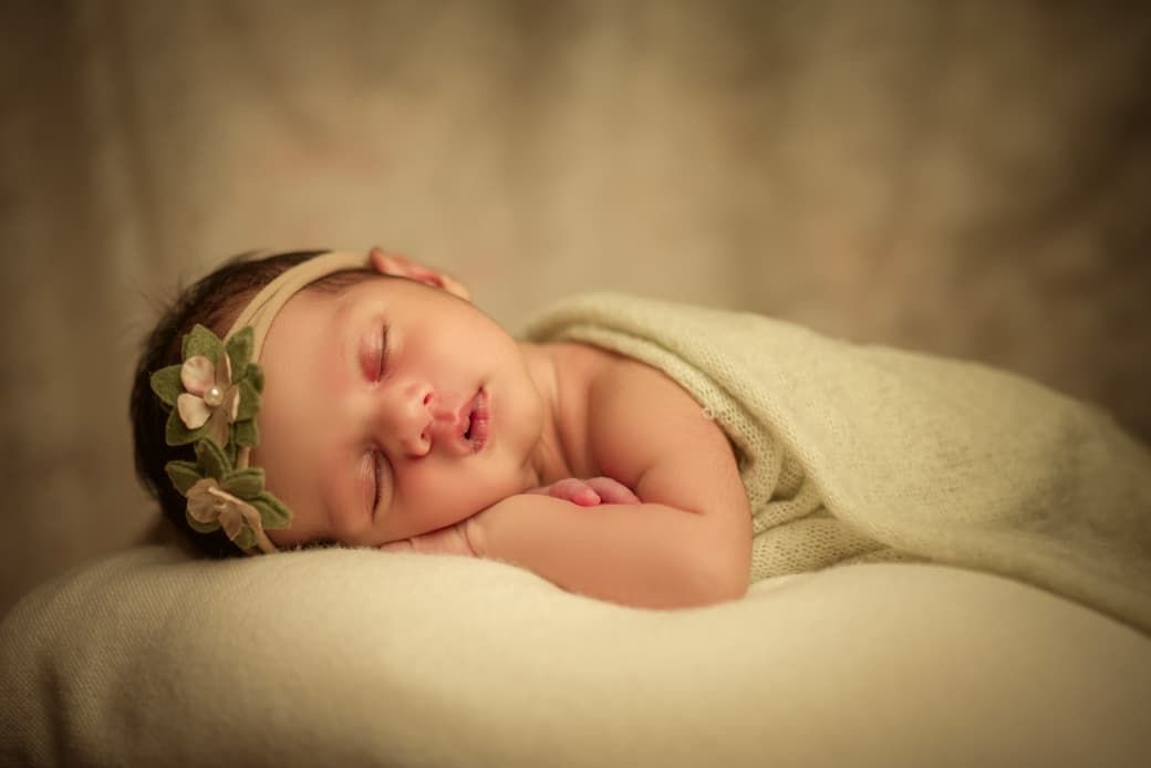 A peacefully sleeping newborn baby wrapped in soft green fabric wearing a delicate floral headband in a studio portrait.
