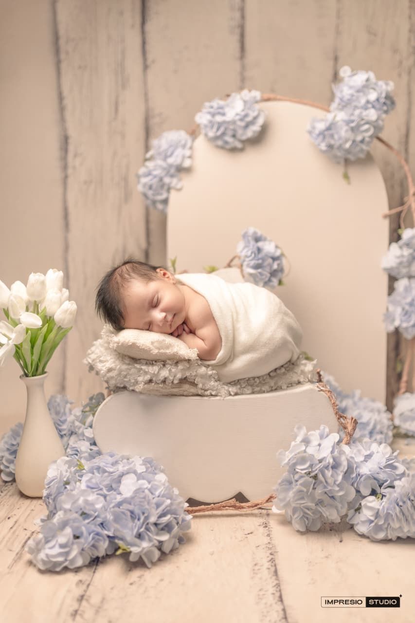 Newborn baby swaddled in white, sleeping peacefully in a decorative box surrounded by blue flowers and white tulips.