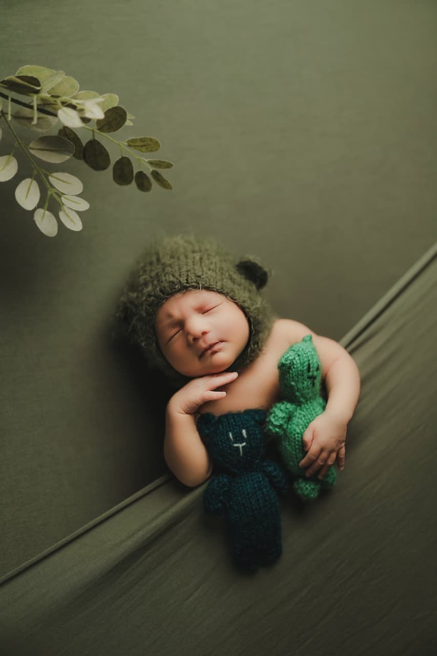 Sleeping newborn baby wearing a green knit bear hat, holding a crocheted dinosaur toy on a green backdrop.