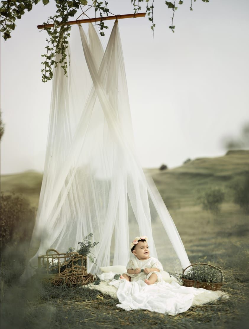 Baby in white dress sits under sheer canopy outdoors on blanket surrounded by wicker baskets in misty field.