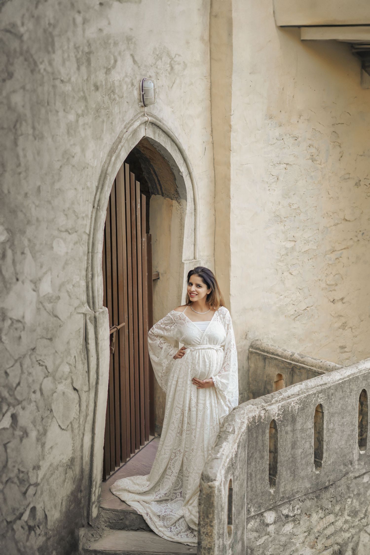 A figure in a flowing white lace dress poses gracefully against an old stone wall with arched doorways.