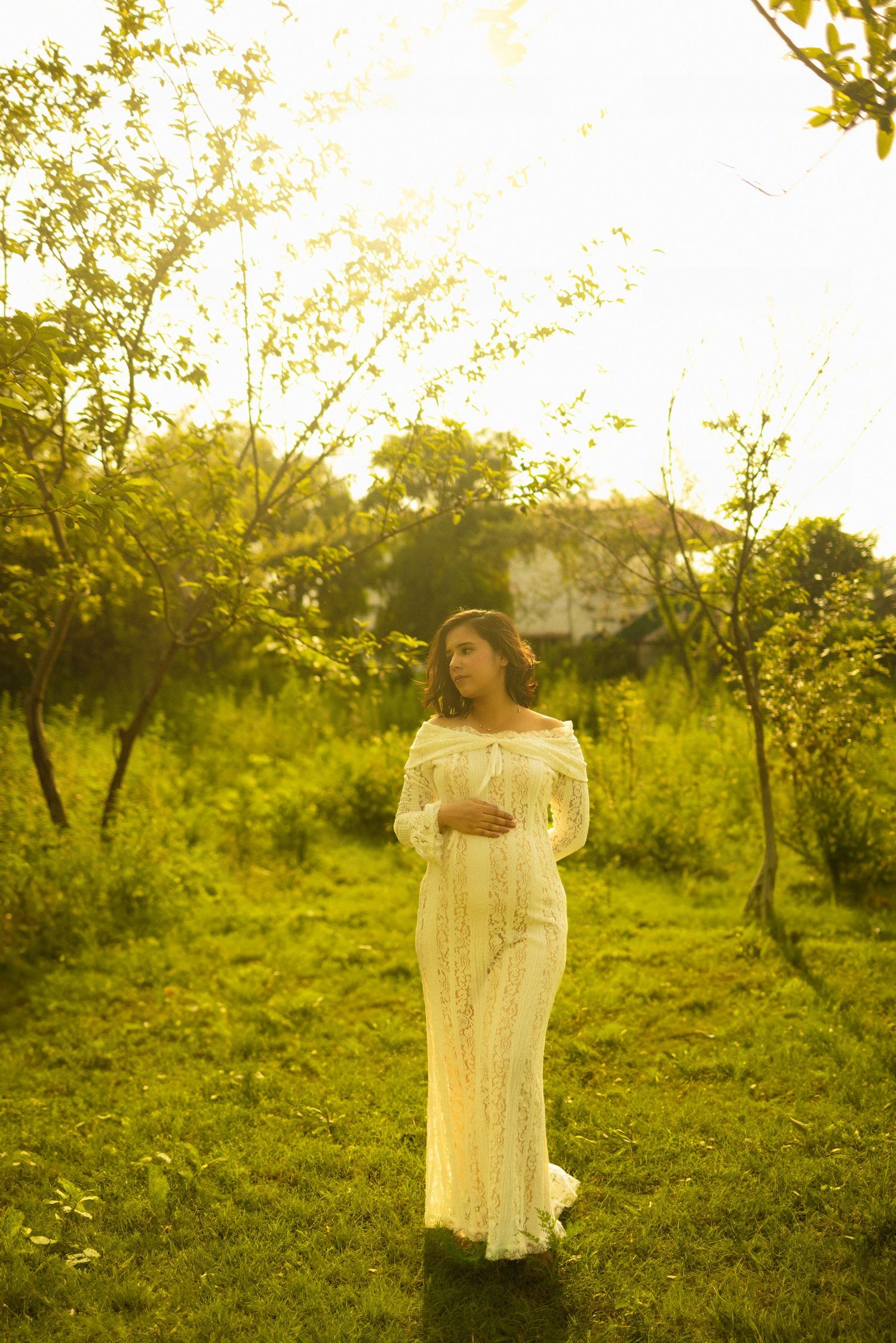 A dreamy maternity portrait set in a sunlit meadow with vintage white lace dress against golden hour lighting and lush greenery.