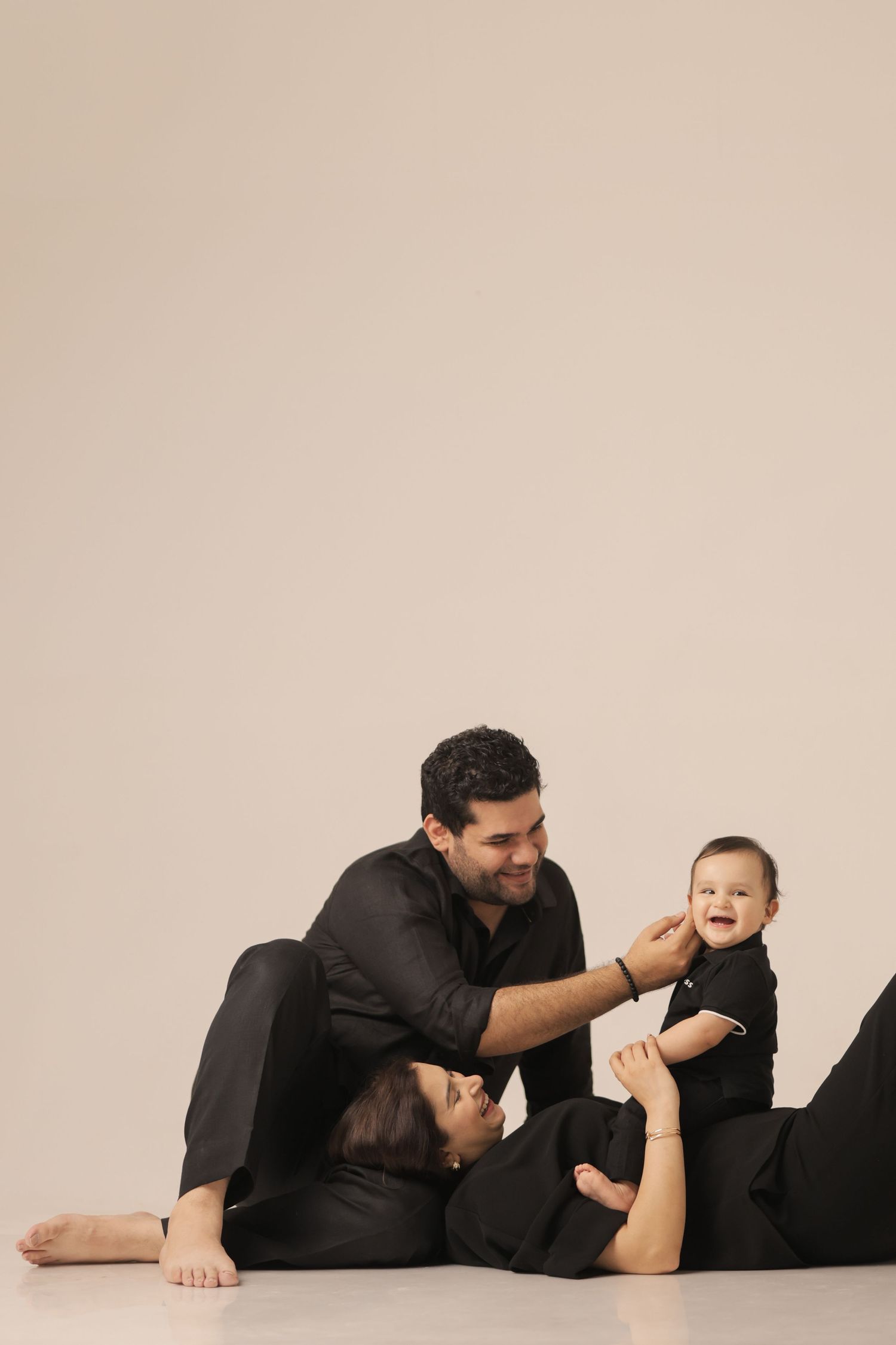 Family in black outfits sitting on floor, parents playing with smiling baby in a studio setting.