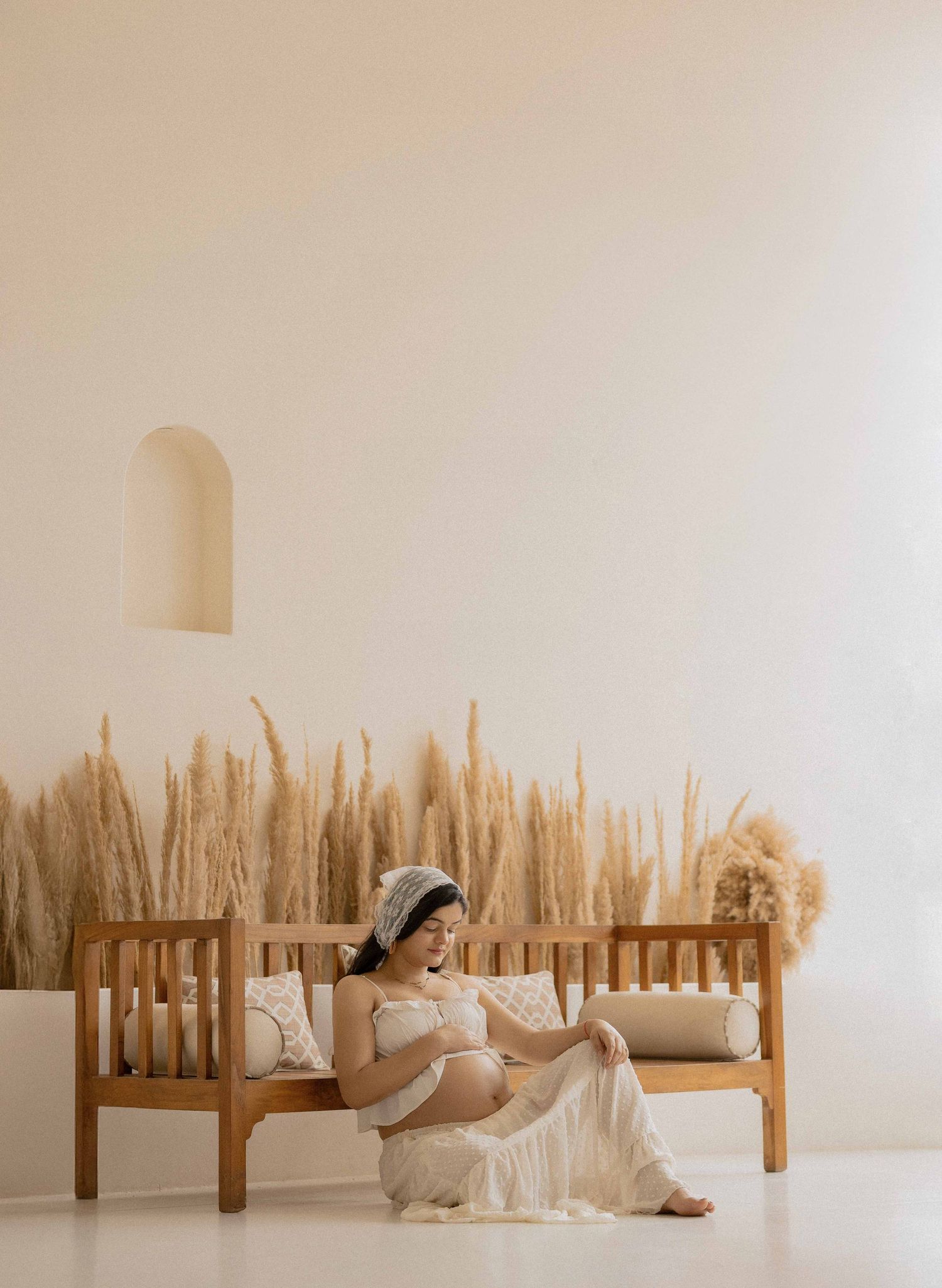 Pregnant woman in white lace dress sitting on wooden bench with pampas grass backdrop in minimalist studio.
