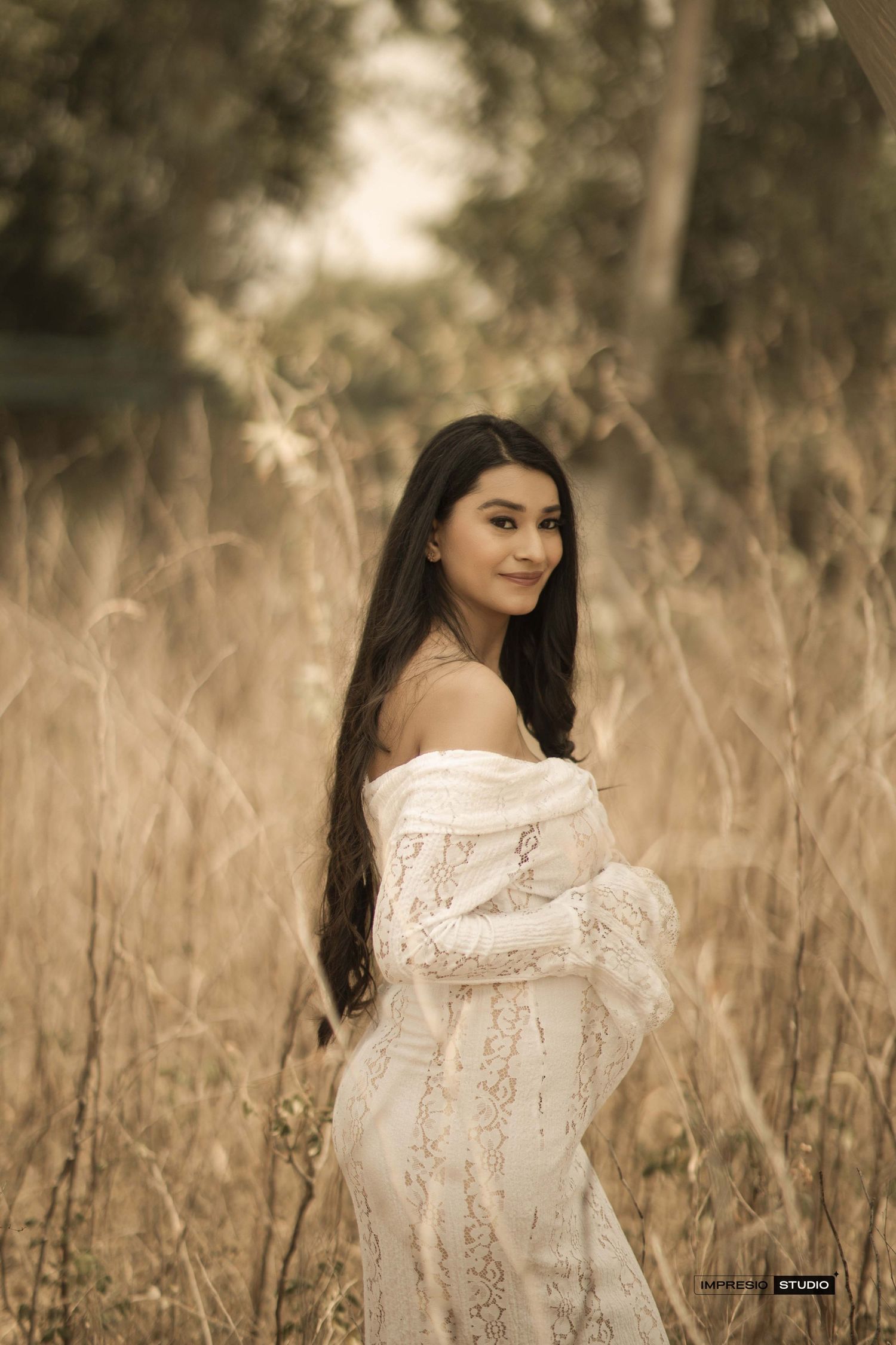Young woman in white off-shoulder lace dress standing in golden field, smiling over shoulder.