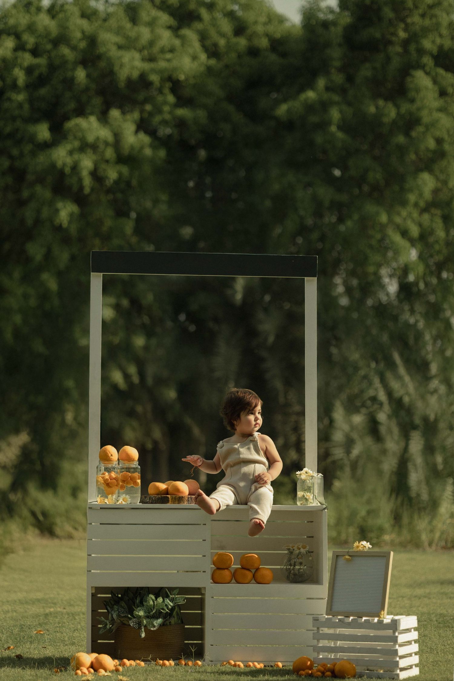 Toddler sitting on white wooden crate stand with oranges and bread rolls outdoors in green park setting.