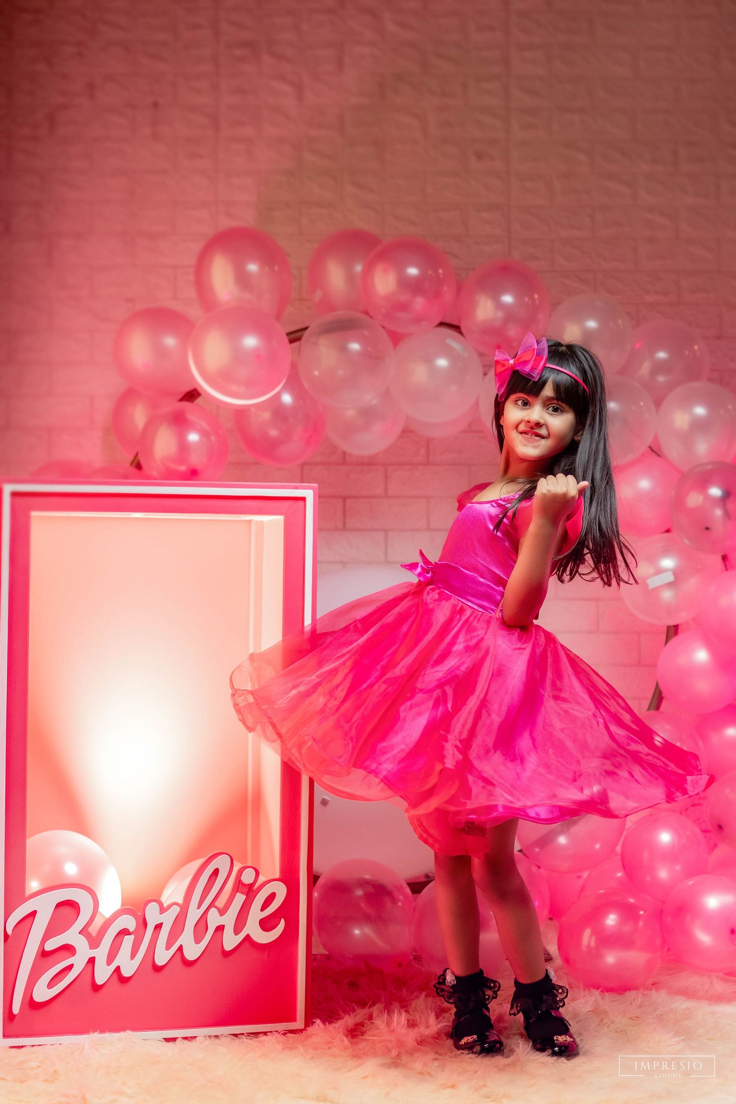 Young girl in pink Barbie dress posing at a Barbie-themed birthday party with pink balloons backdrop.