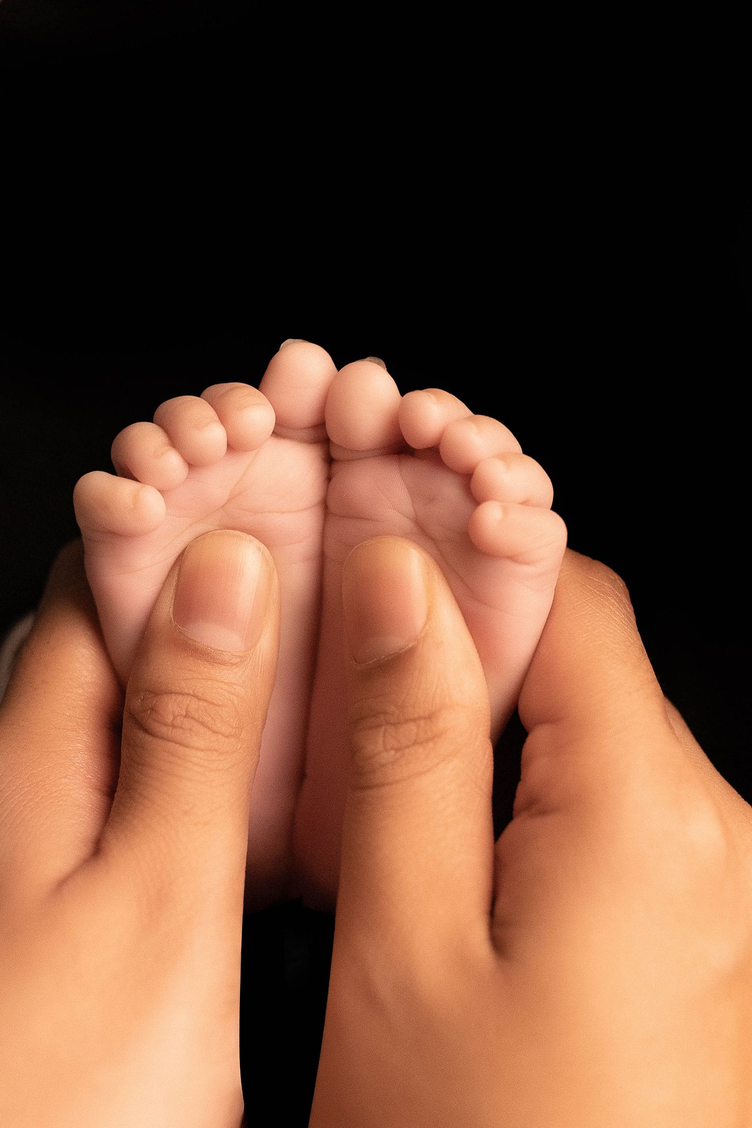 Adult hands gently holding tiny newborn baby feet against a black background, showcasing delicate infant toes.