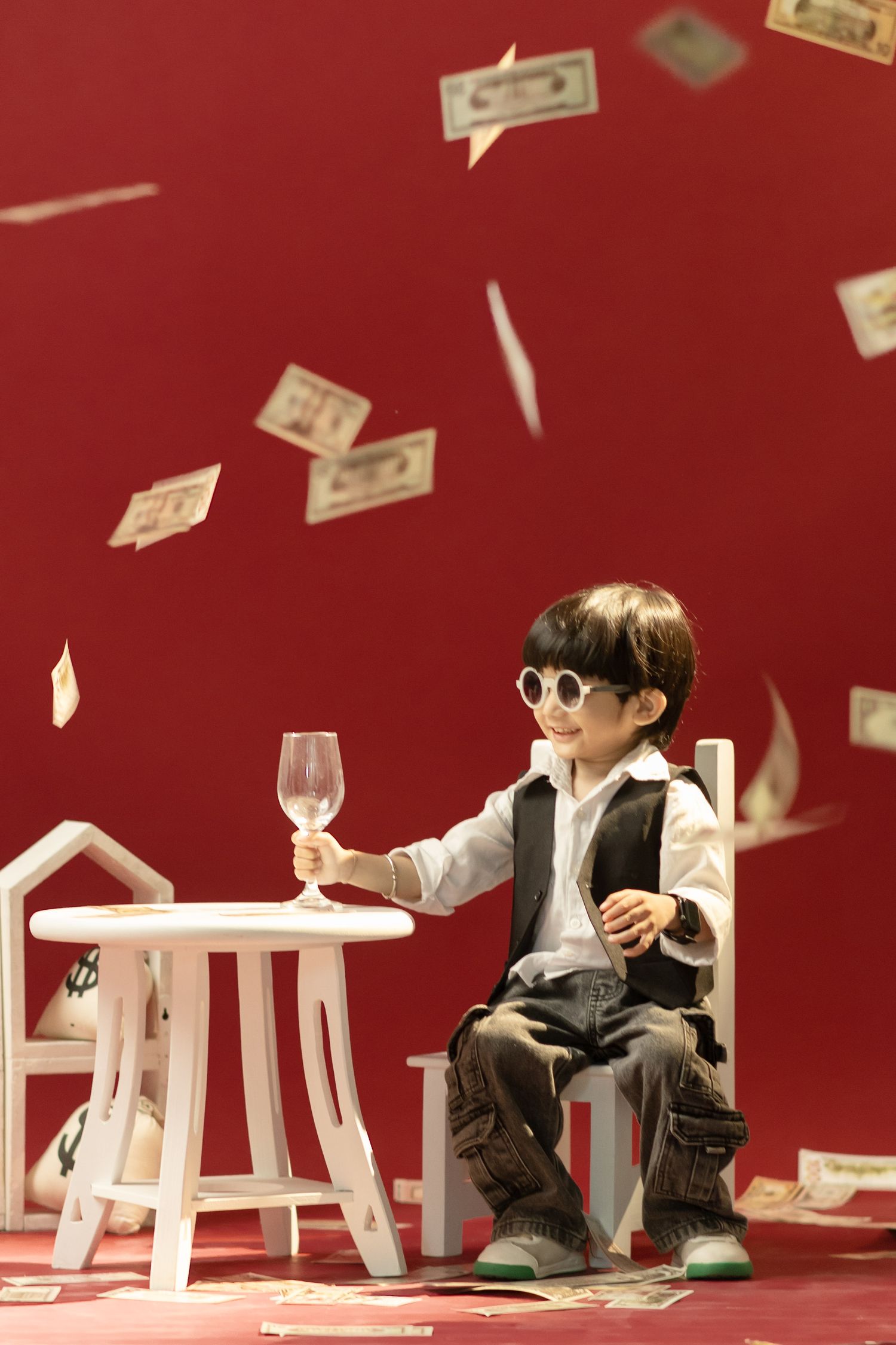 Young boy in sunglasses holding a wine glass at a white table with money raining down on red background.