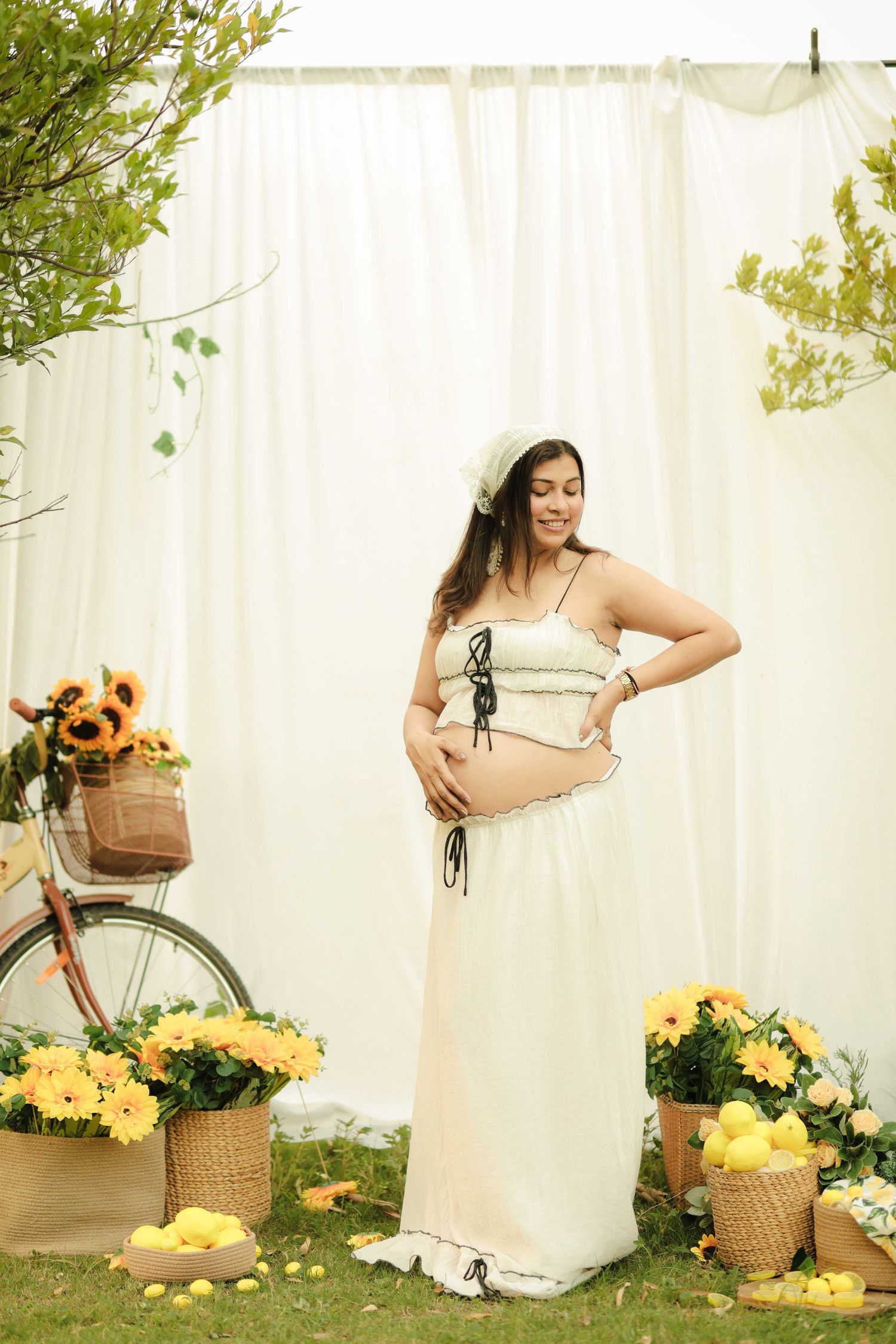 Pregnant woman in white crop top and maxi skirt holding belly, surrounded by yellow flowers and sunflowers outdoors.