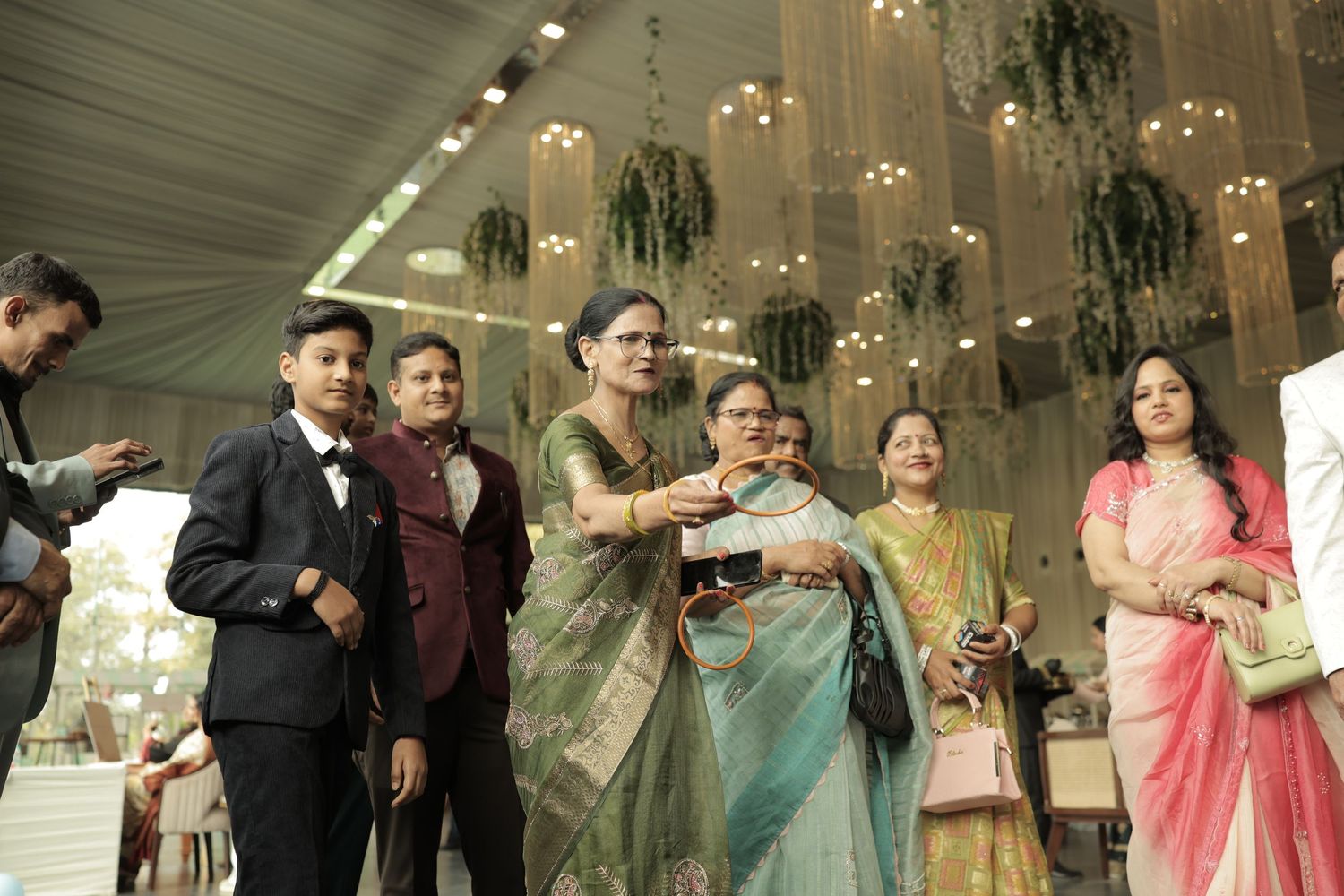 Family gathering at an elegant Indian wedding reception with guests in traditional attire under decorative chandelier.