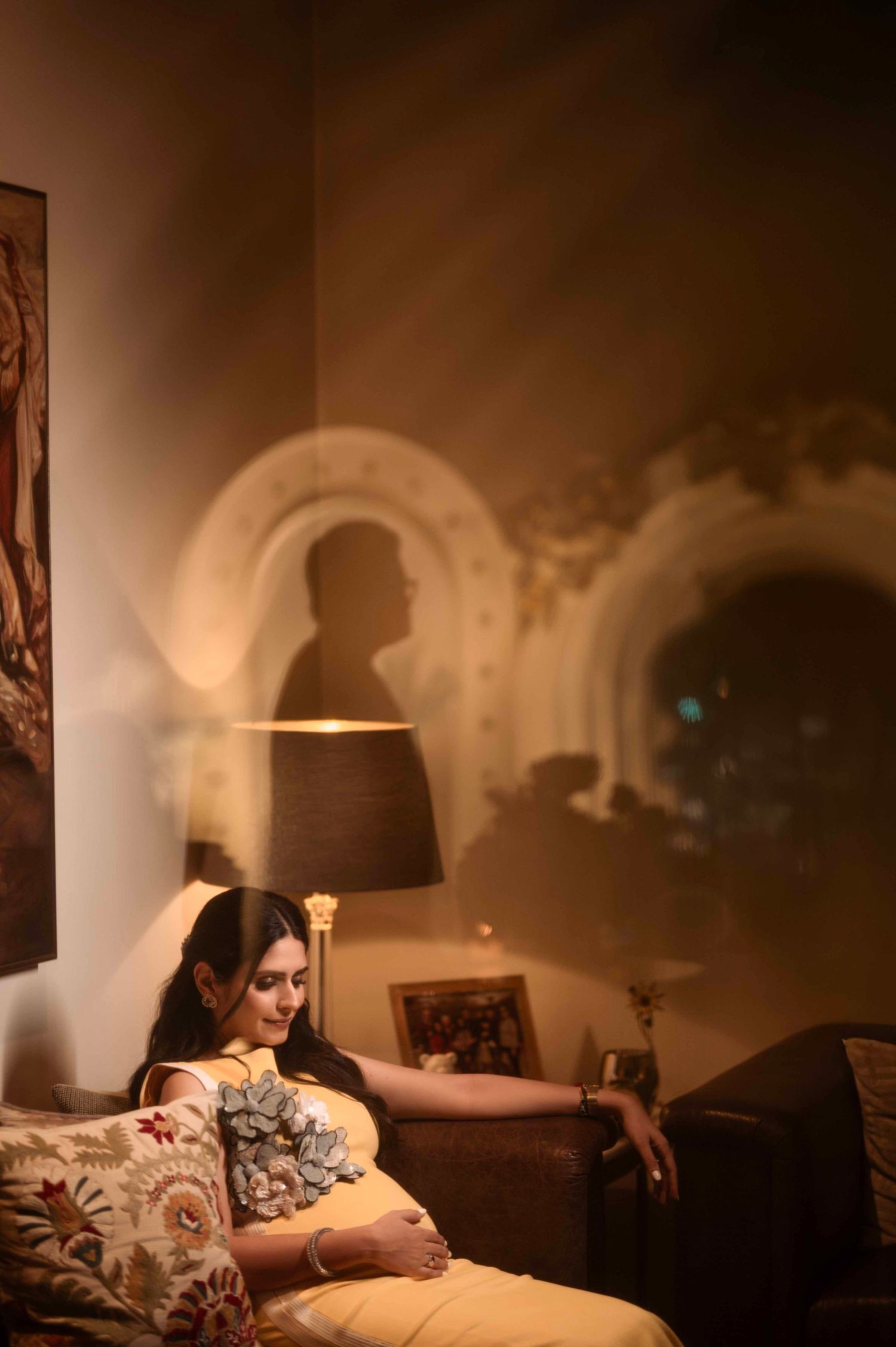 Pregnant woman in elegant attire resting on sofa in warmly lit room with artistic shadow on wall.