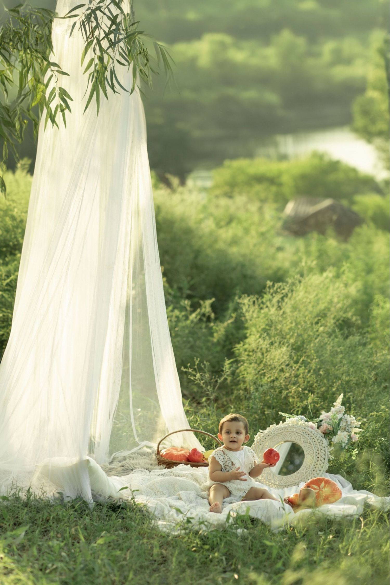 Baby sitting on white blanket outdoors under sheer canopy with fruits and mirror in lush green garden setting.