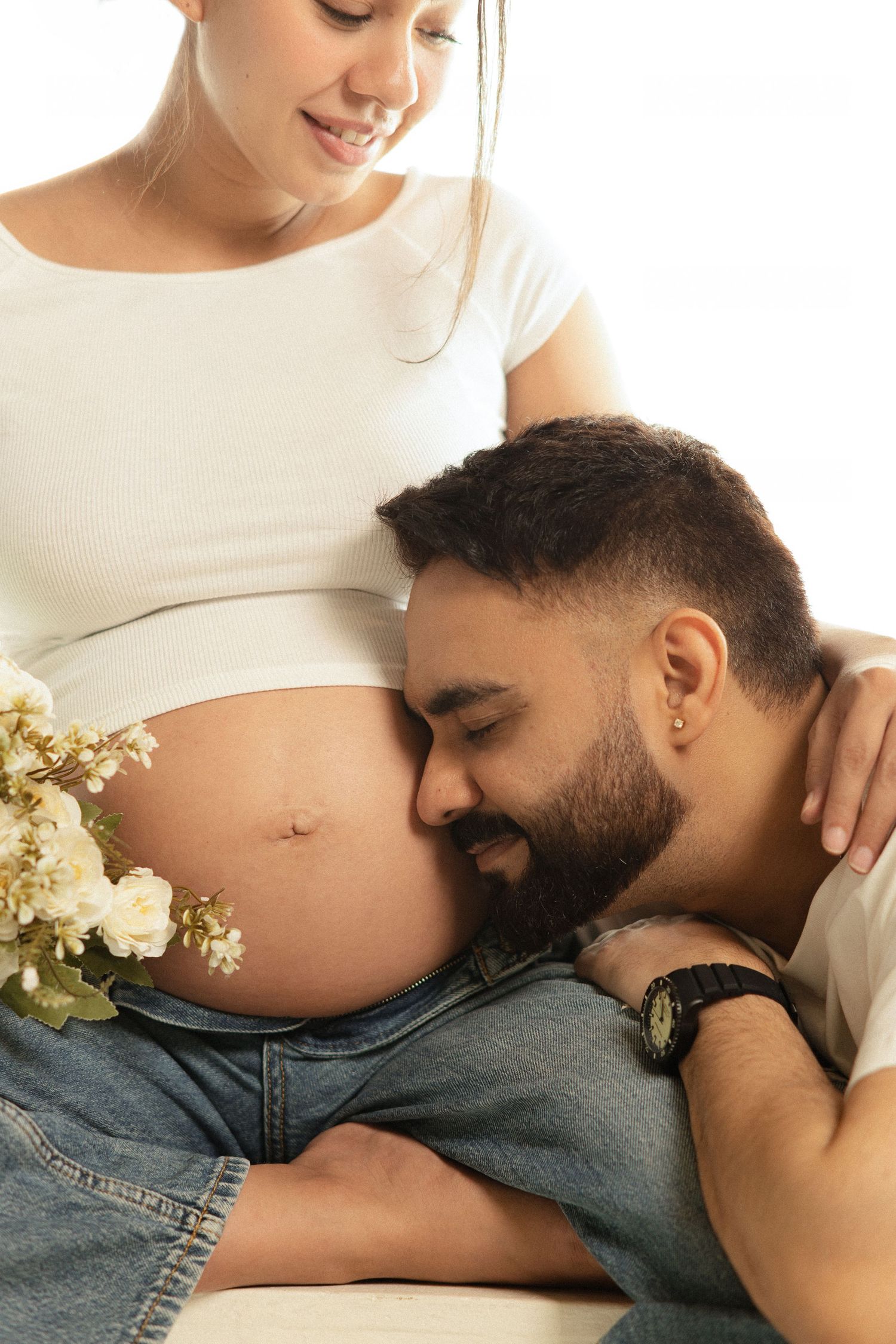 A bearded man rests his head on a pregnant woman's belly, smiling, while she holds white flowers.