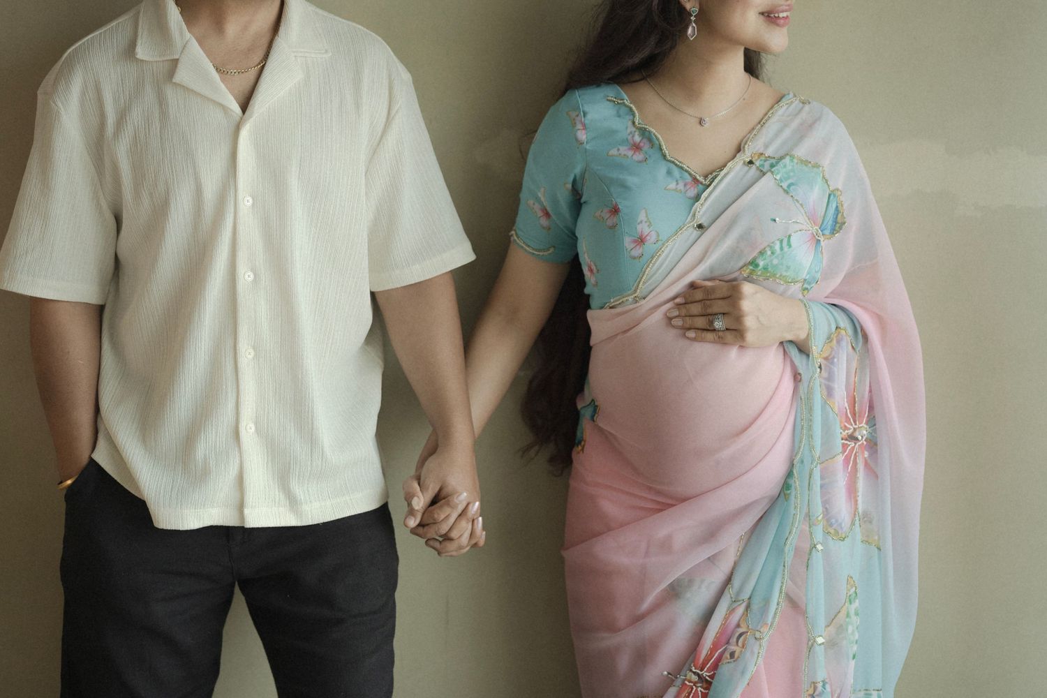 Couple holding hands, man in white shirt and woman in pink and blue floral saree, maternity photo.