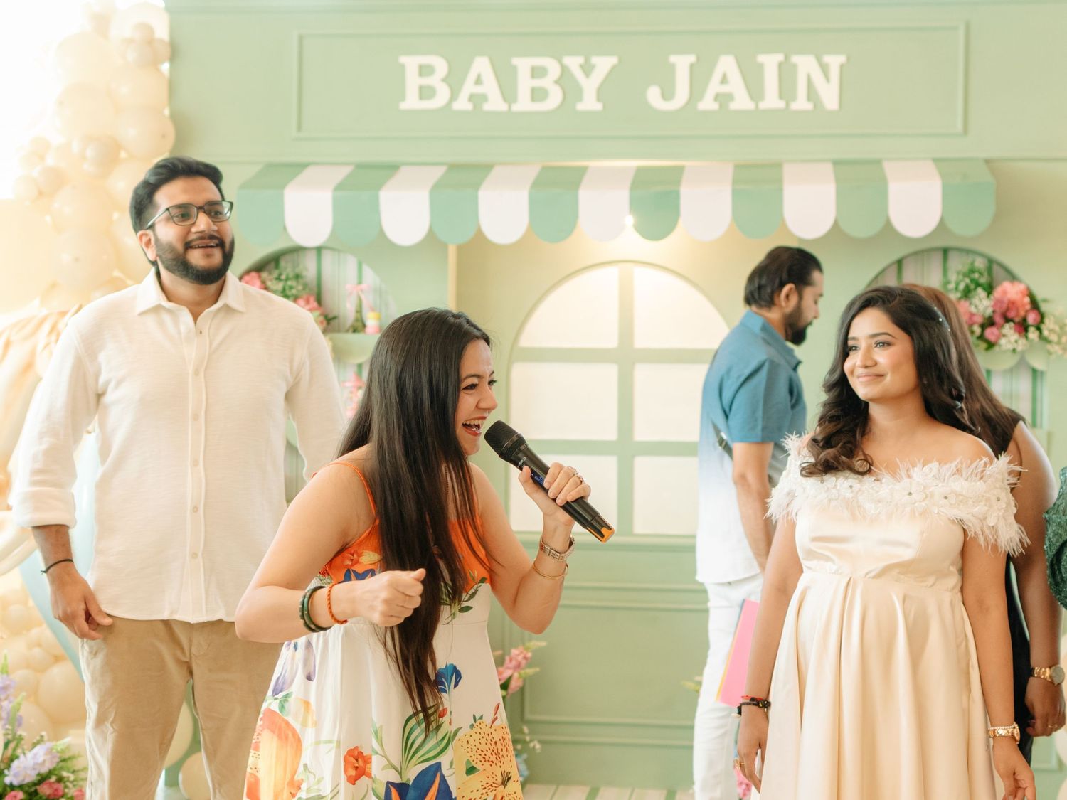 Women celebrate at a Baby Jain baby shower event, one holding a microphone, with a decorative green storefront backdrop.