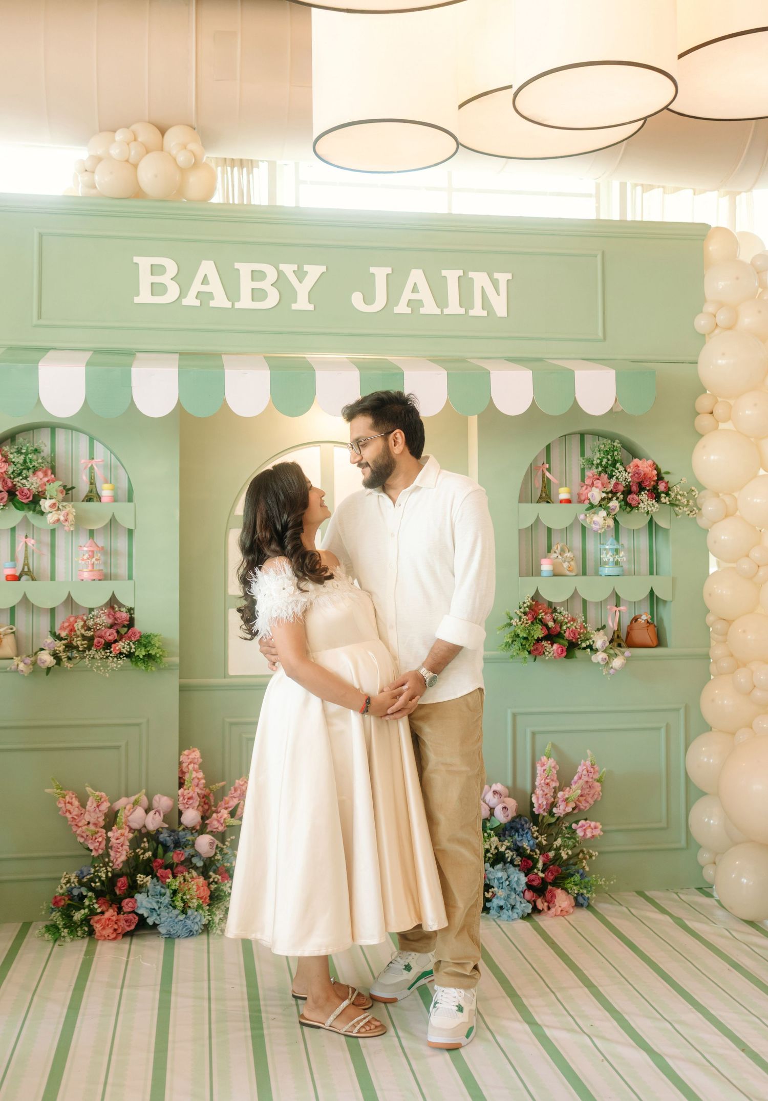 Couple poses at a sage green Baby Jain baby shower backdrop with floral decor and balloon arch.