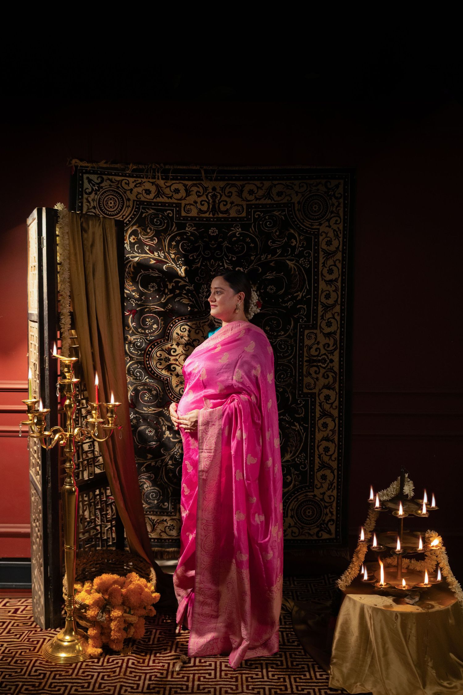 Person wearing vibrant pink sari stands in elegant room with ornate tapestry, candles and traditional decor.