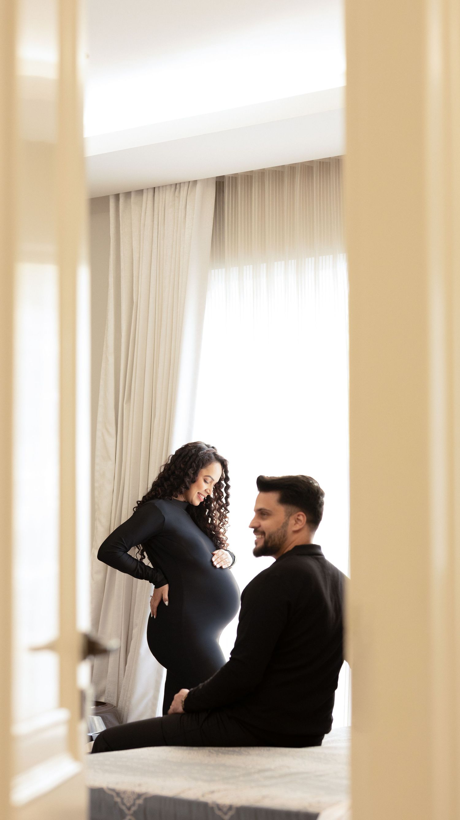 A tender moment between an expectant couple by a bright window with white curtains in a softly lit room.