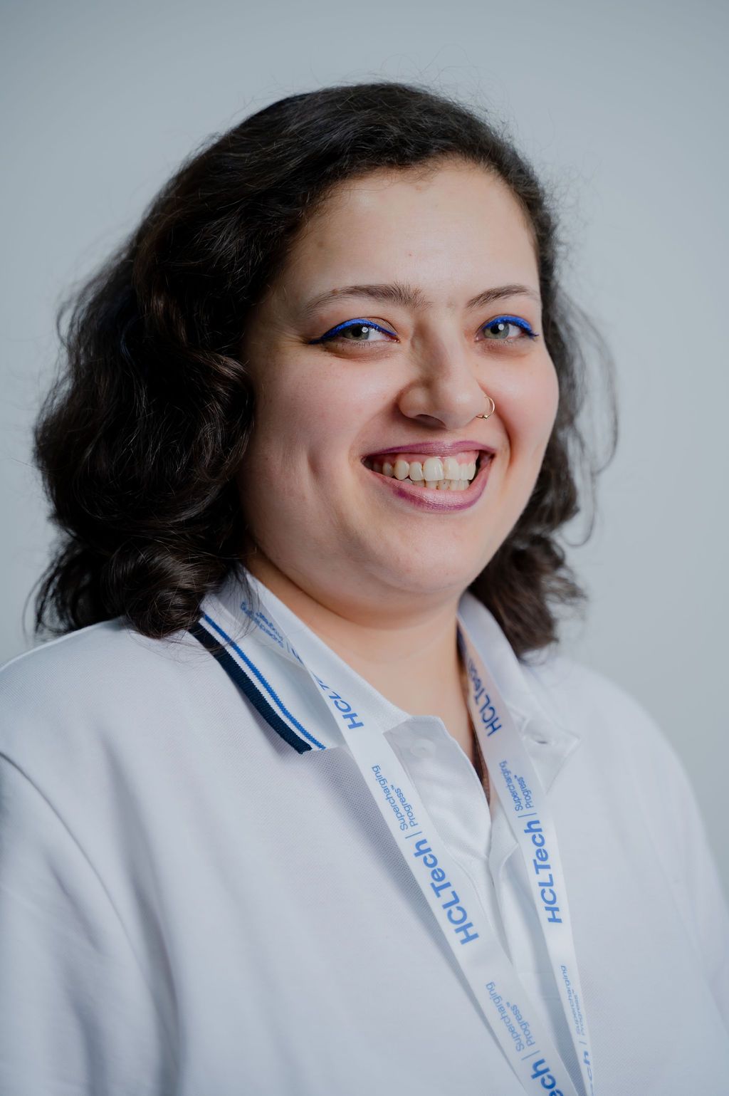 Smiling woman with blue eye makeup wearing a white top and HCLTech lanyard against a gray background.