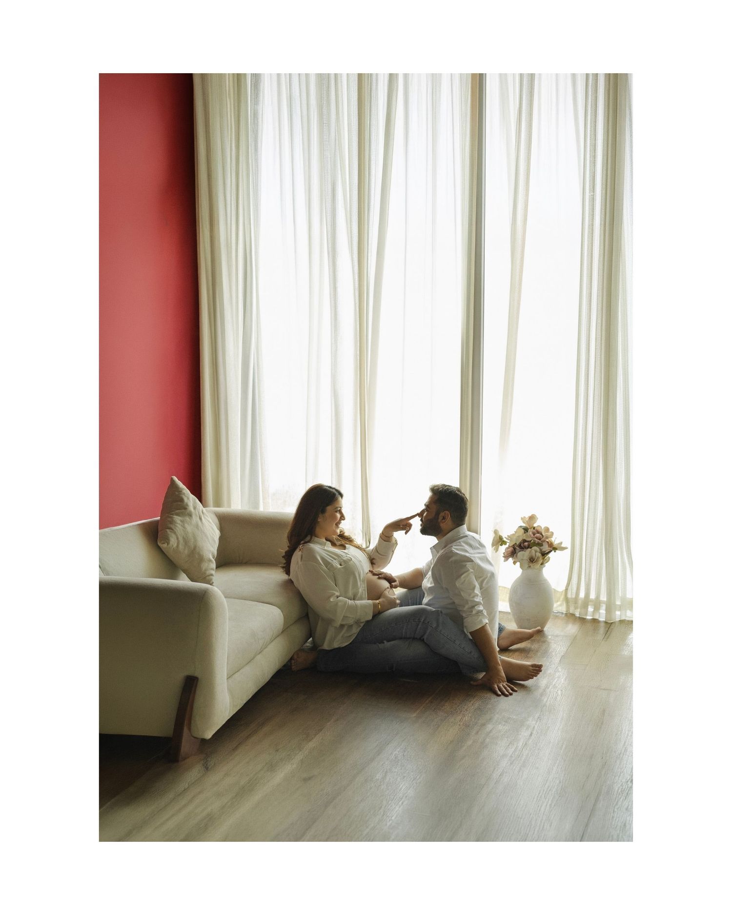 Couple sitting on floor by sofa near bright curtains, sharing a romantic moment in a modern living room.