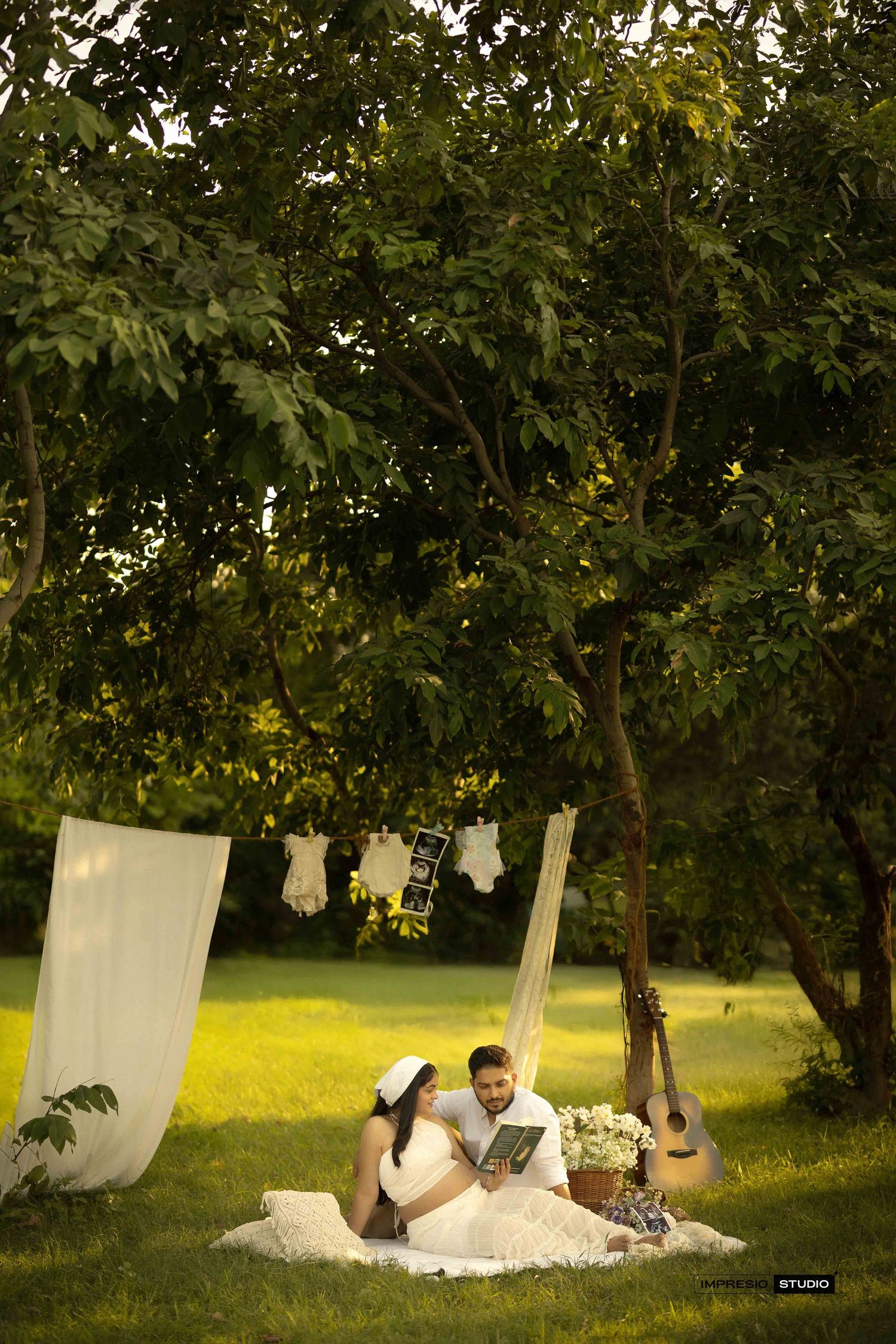 Couple sitting on grass under trees during maternity photoshoot with baby clothes hanging and guitar nearby.