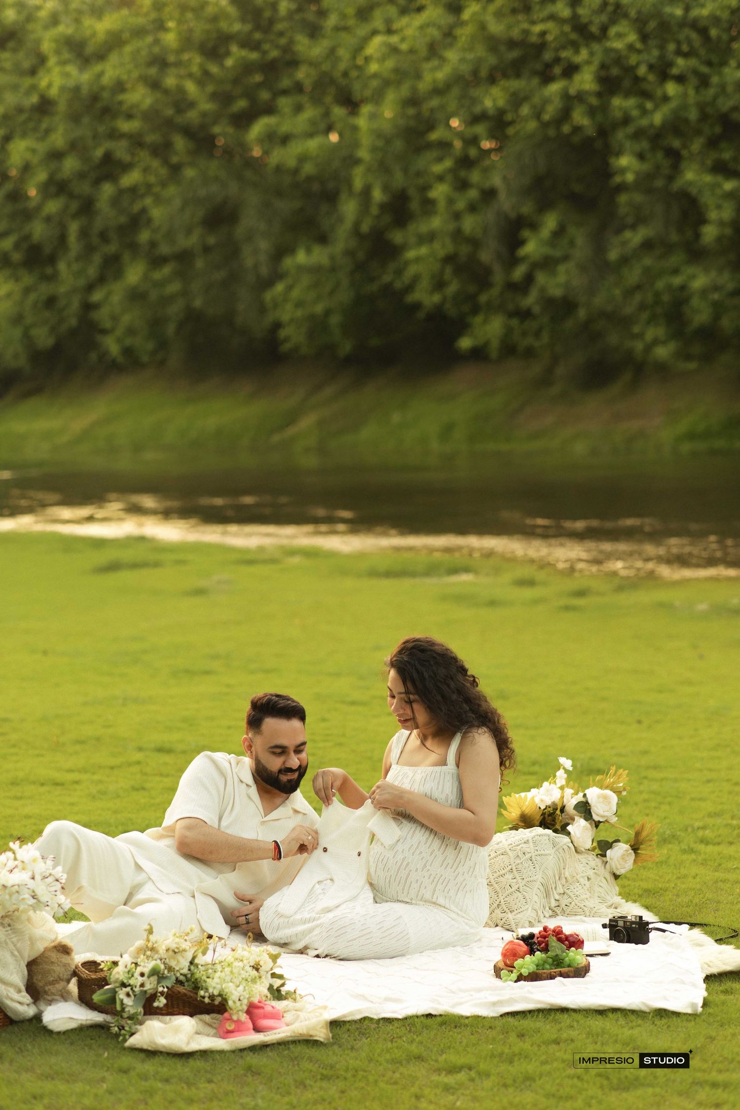 A couple in white outfits enjoy a romantic outdoor picnic on a green lawn beside a serene river.