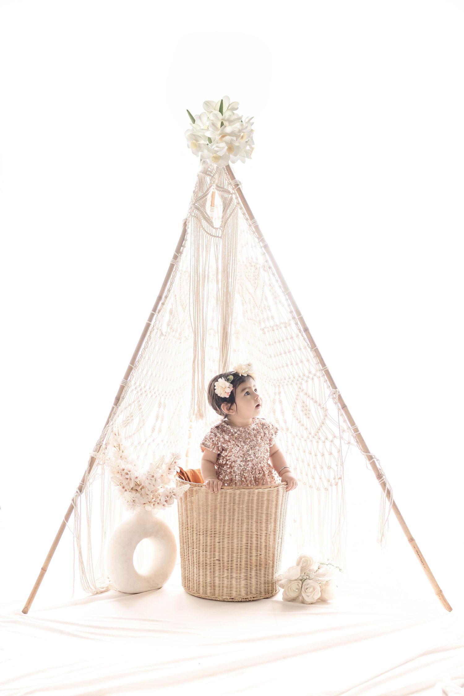 Baby girl in pink outfit sits in wicker basket under boho macrame teepee with white flowers and decor.