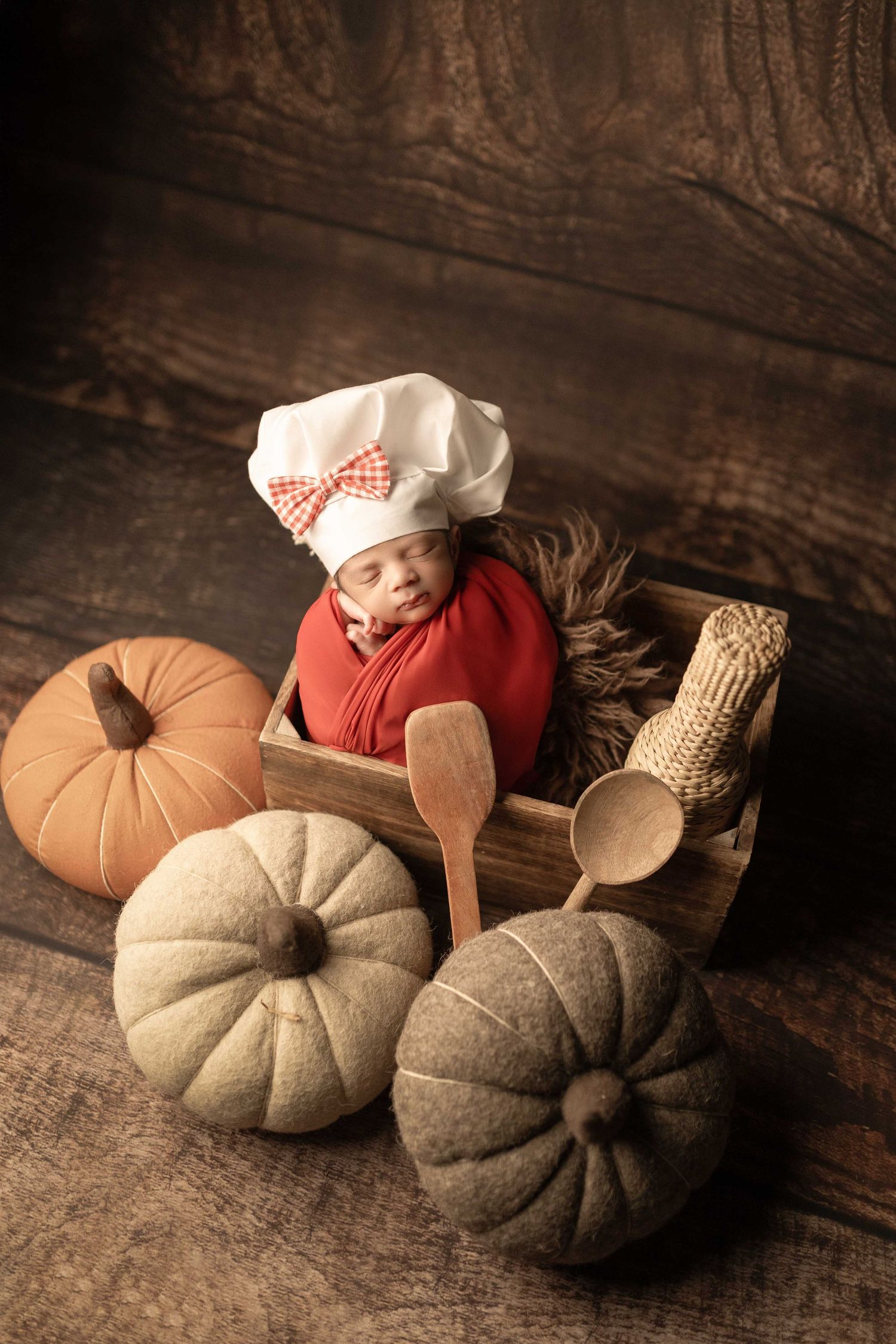 Newborn baby dressed as a chef in a wooden crate surrounded by decorative pumpkins and wooden spoons.